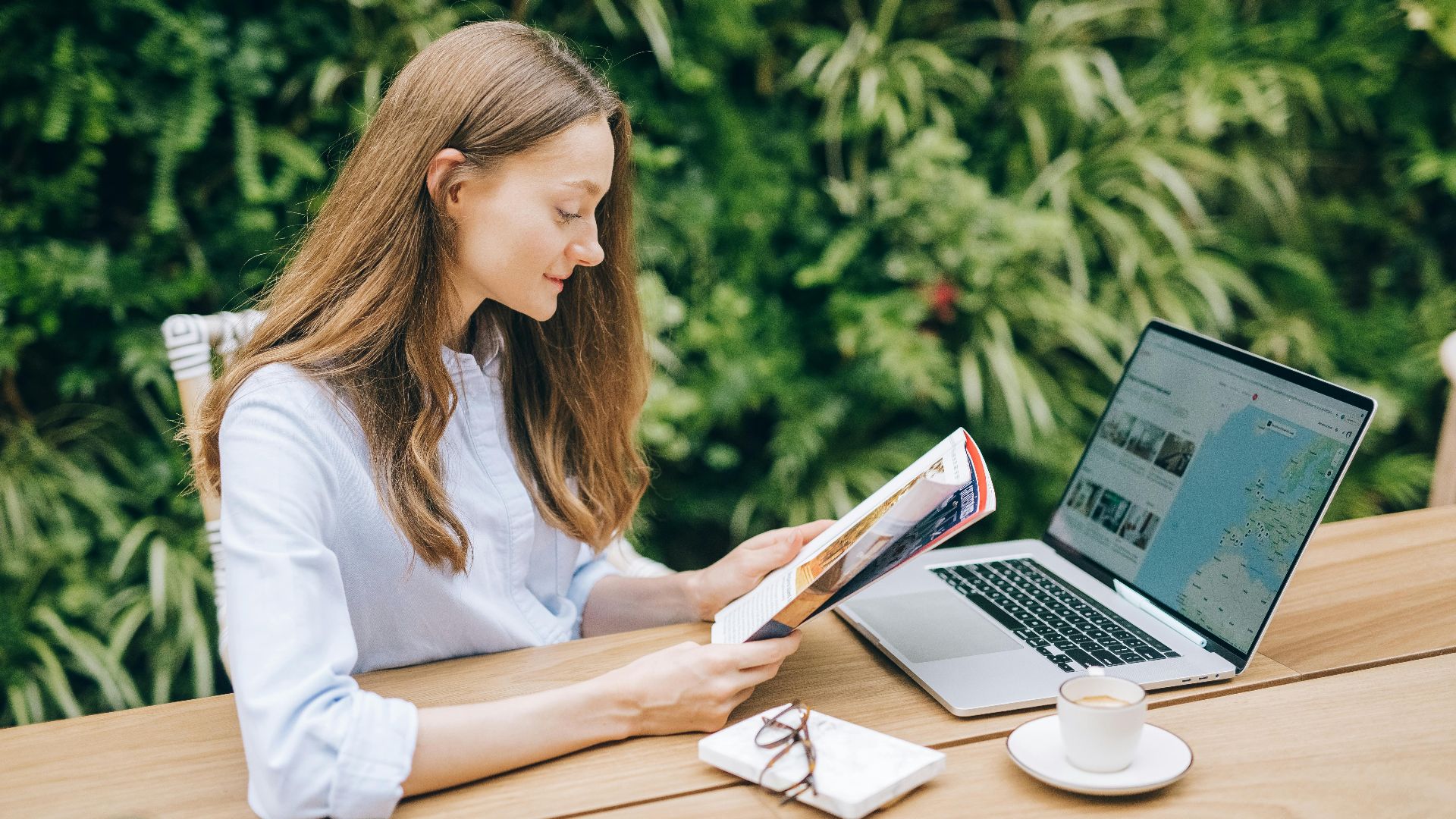 Caucasian woman reading a magazine at a wooden table with a laptop, outdoors.
