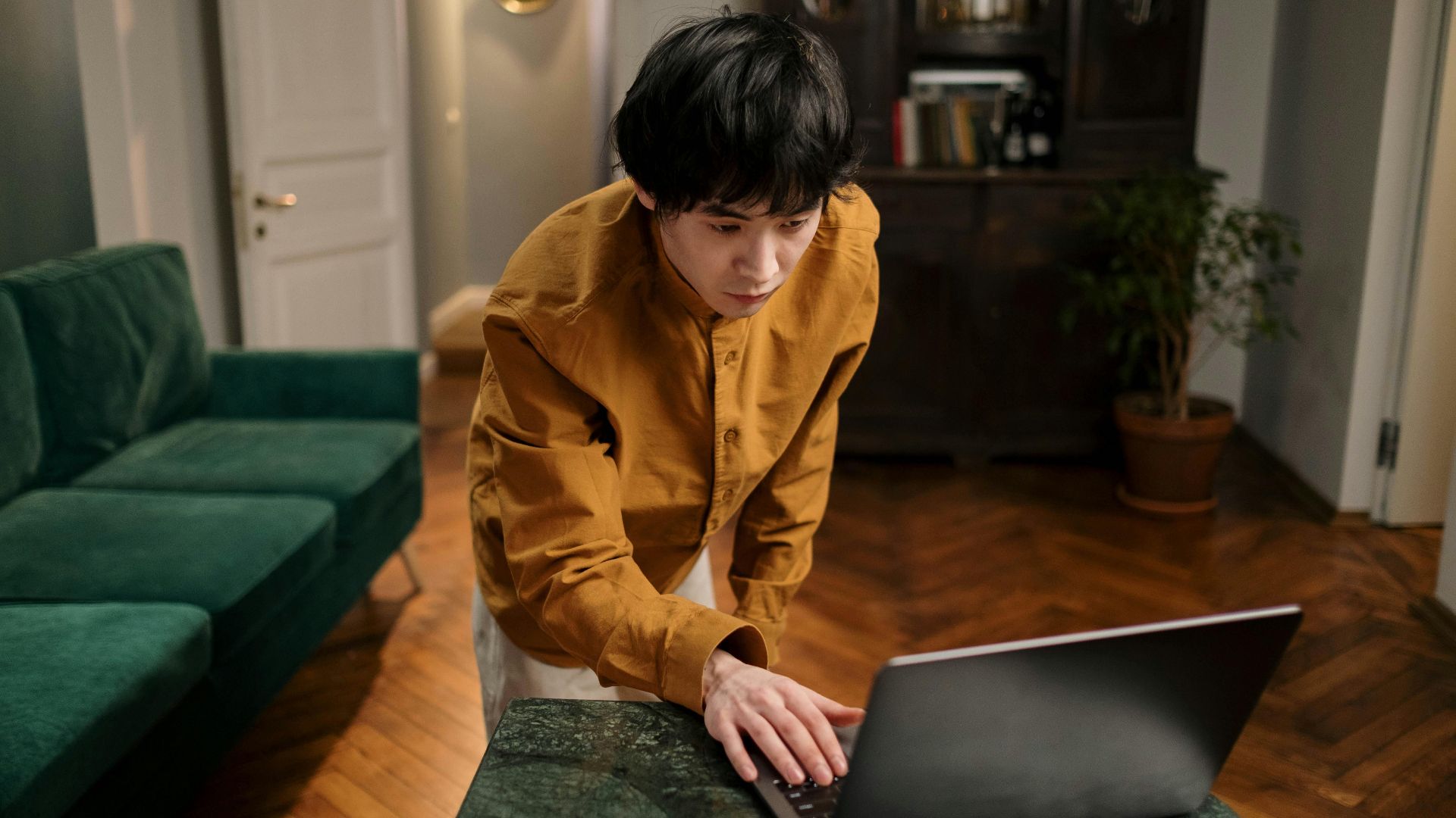 A man standing at a laptop in a stylish living room. Ideal for themes of home office or online learning.