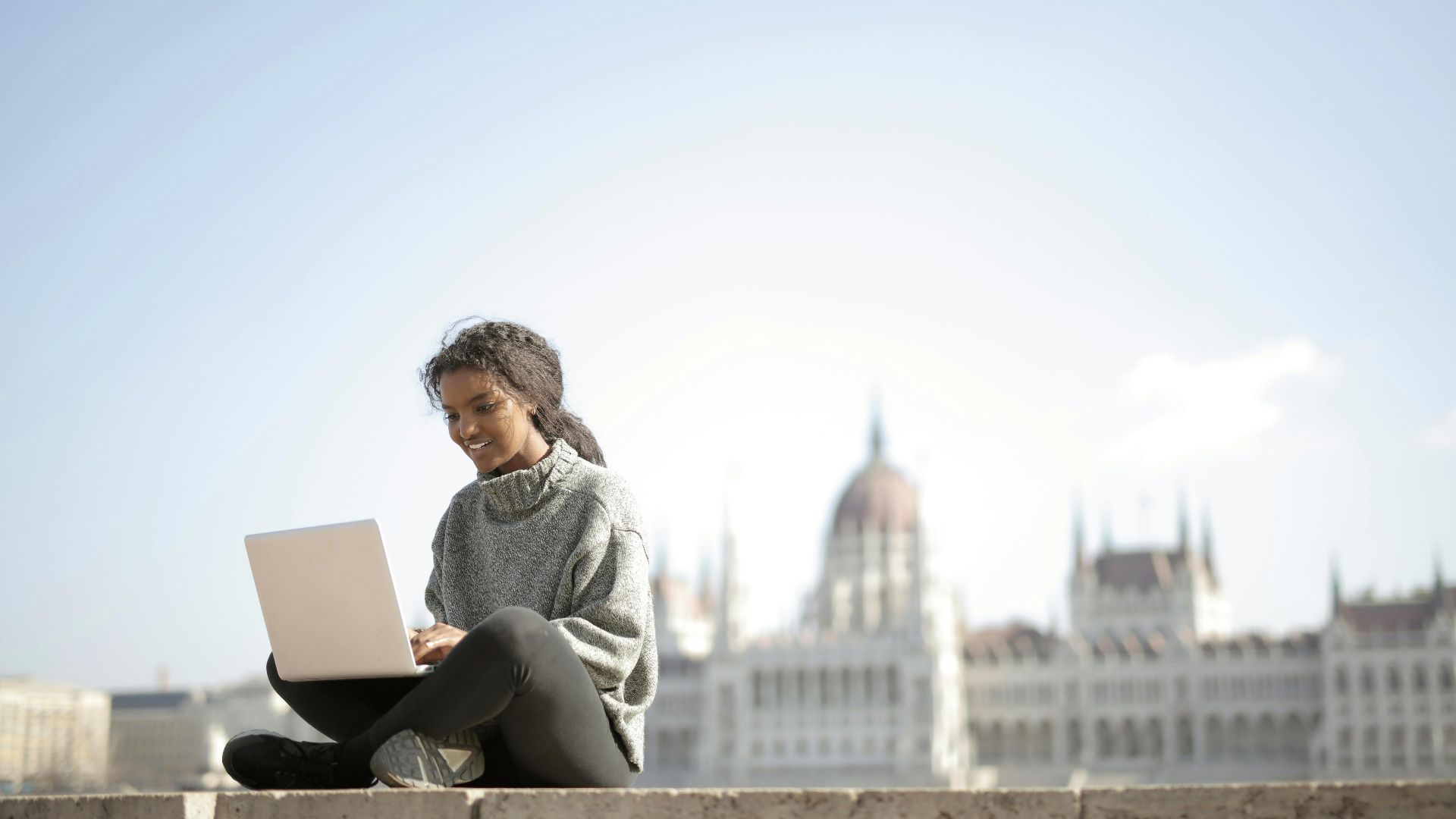Woman working on laptop in front of Budapest's Parliament Building, enjoying outdoor remote work.