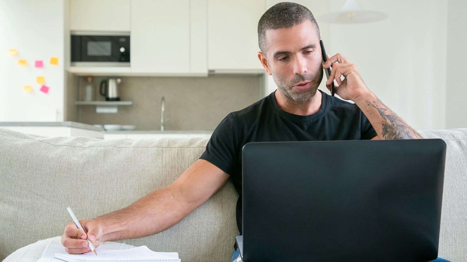 Man working from home using a laptop and smartphone while taking notes in a modern kitchen setting.