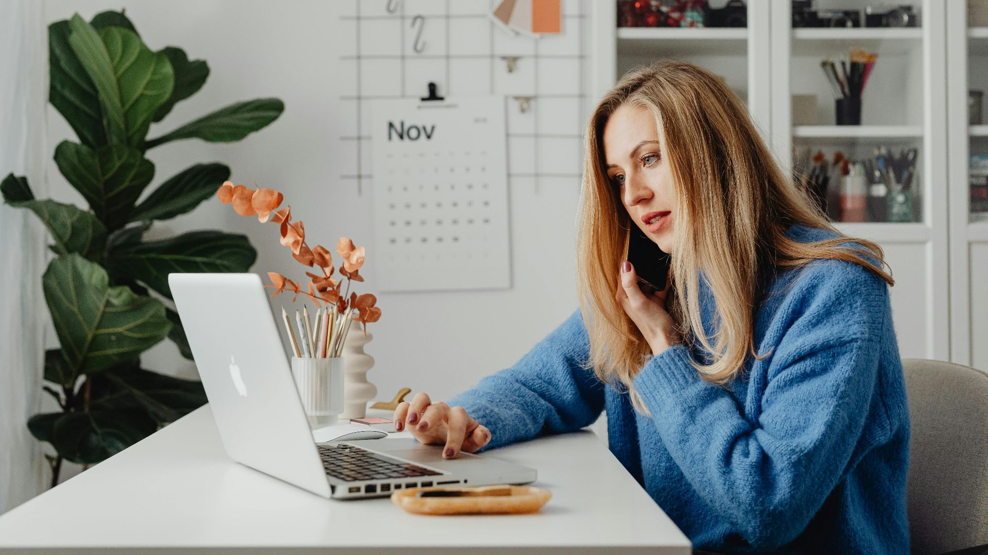 Woman multitasking with laptop and phone in a home office setting.