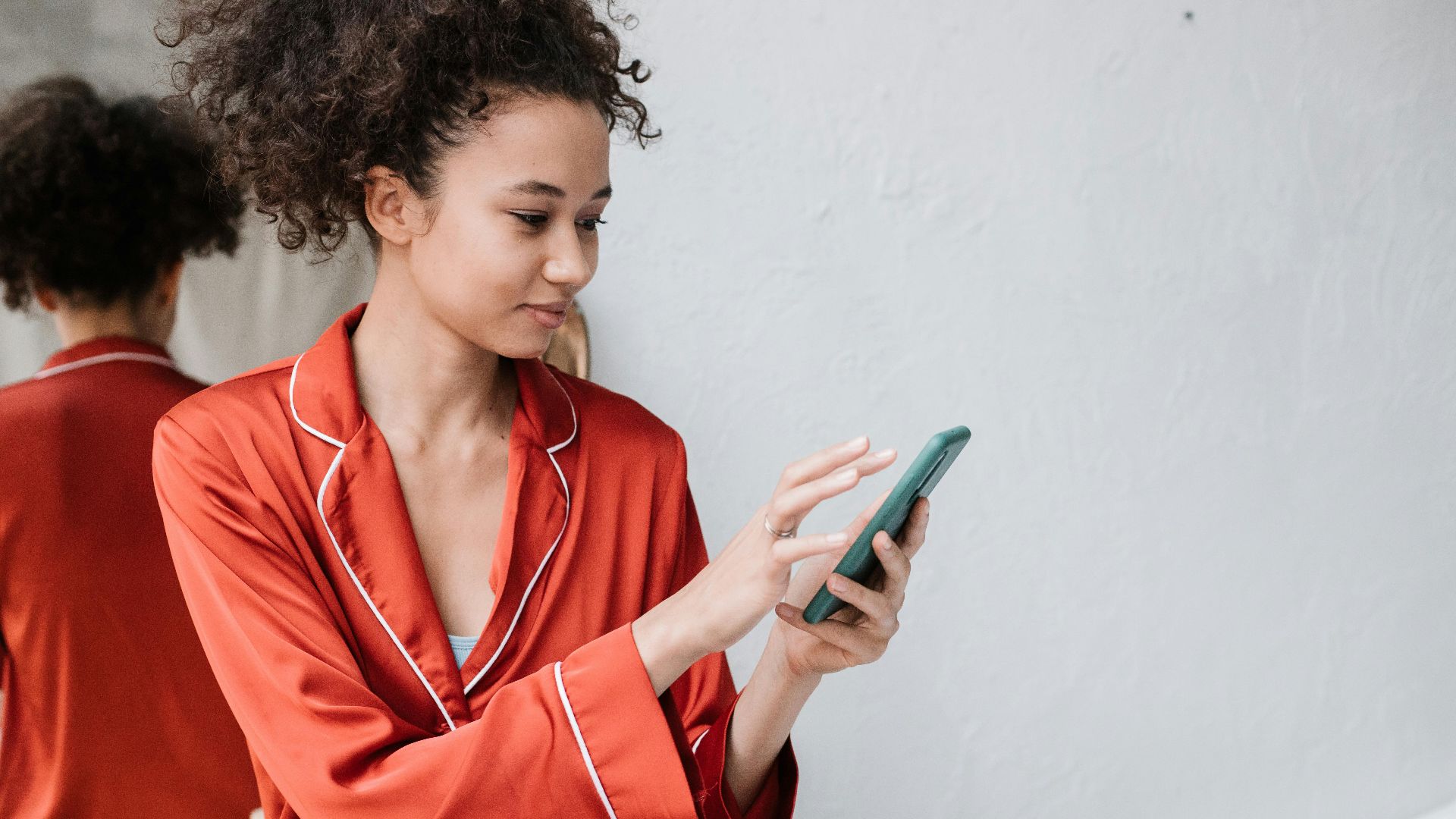 Young woman in red pajamas using smartphone indoors with a mirror reflection, capturing a casual moment.