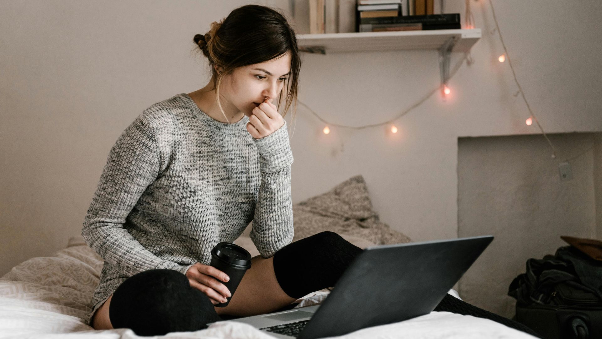 A woman in a gray sweater sits on her bed with a laptop and coffee, focused on work or study in a cozy bedroom.