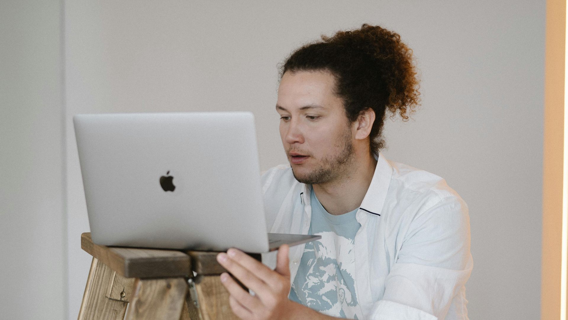 A man using a laptop while standing on a wooden ladder in a minimal indoor setting.