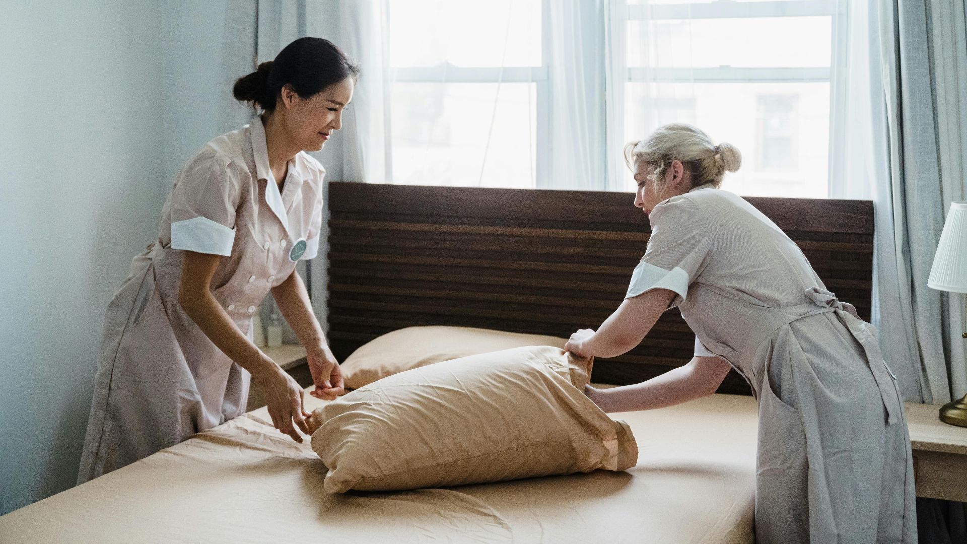 Two housekeepers in uniform collaborate while making a bed in a bright, peaceful bedroom.