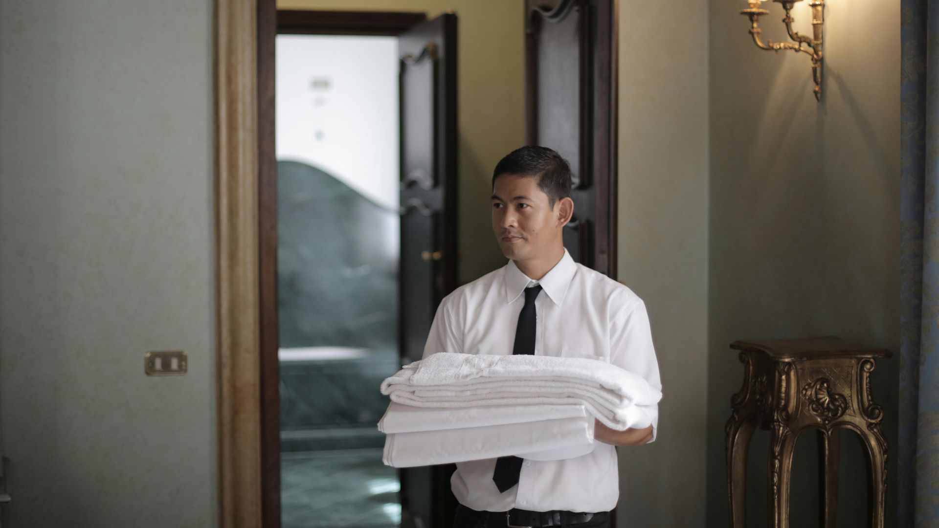 Young male housekeeper carrying stack of white bed sheets while entering bedroom in hotel