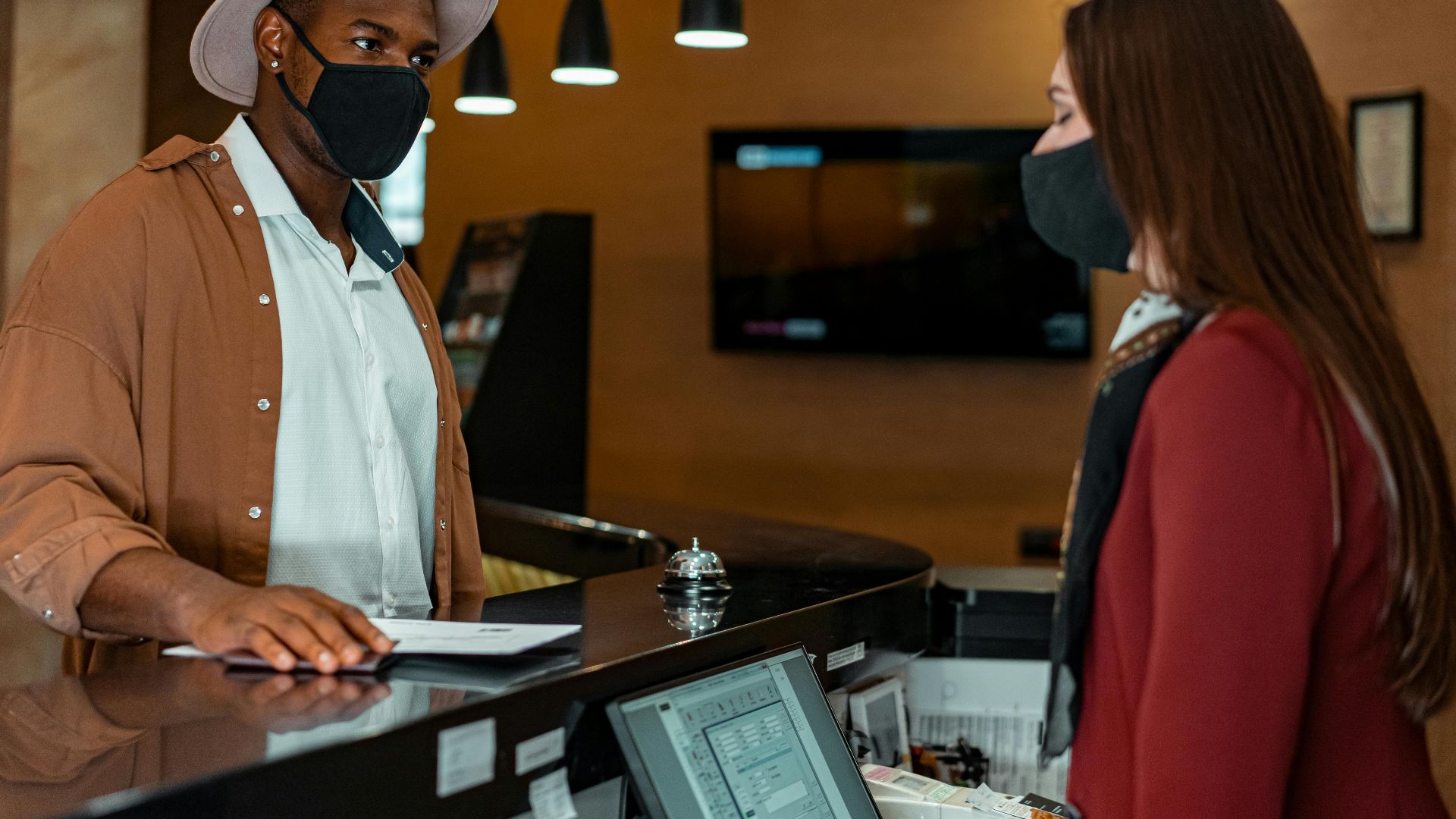 A man and woman in face masks checking in at a hotel reception, emphasizing safety protocols.