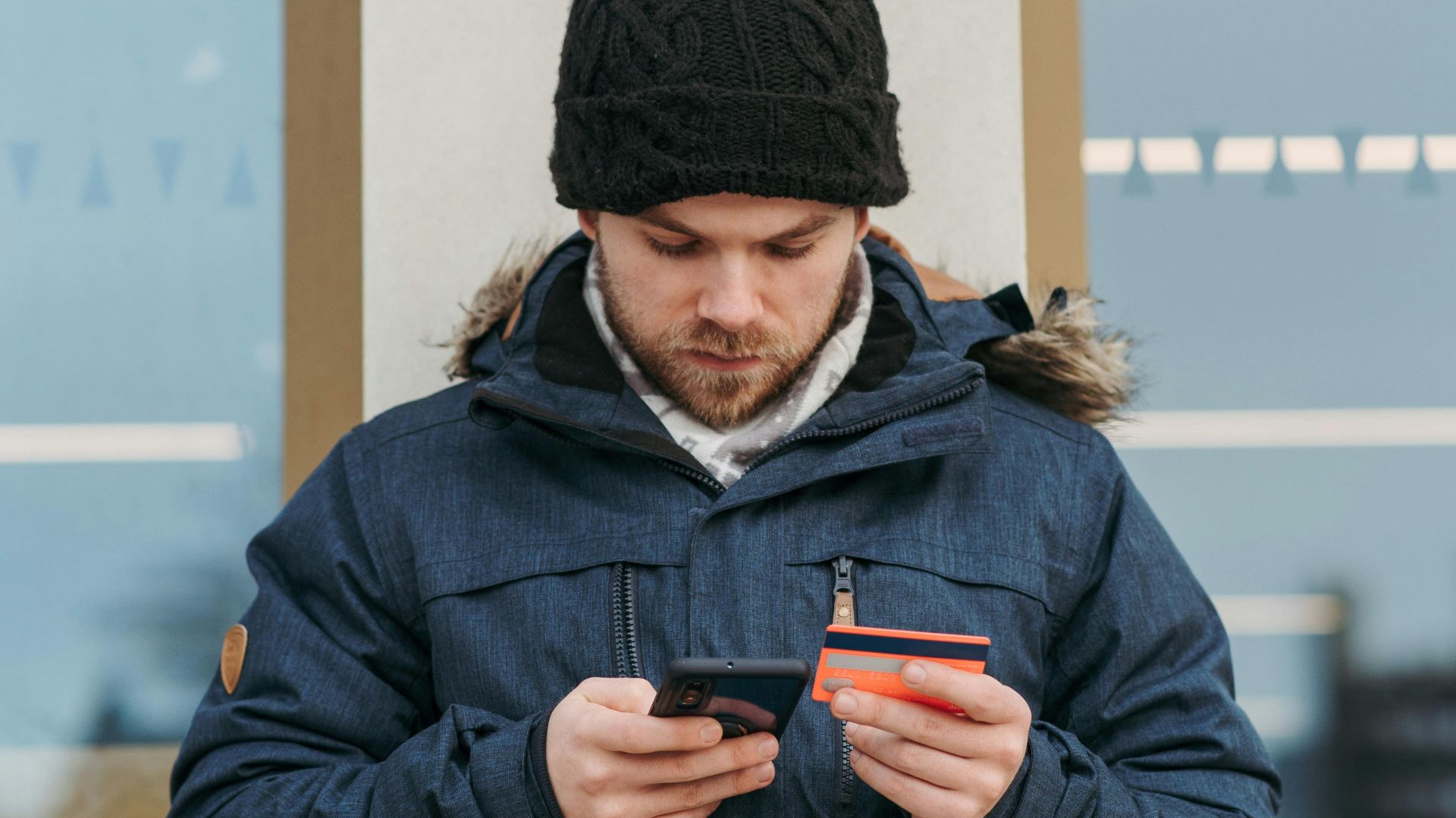Serious young guy in warm clothes and hat standing on city street and browsing smartphone while making online payment with credit card near modern building
