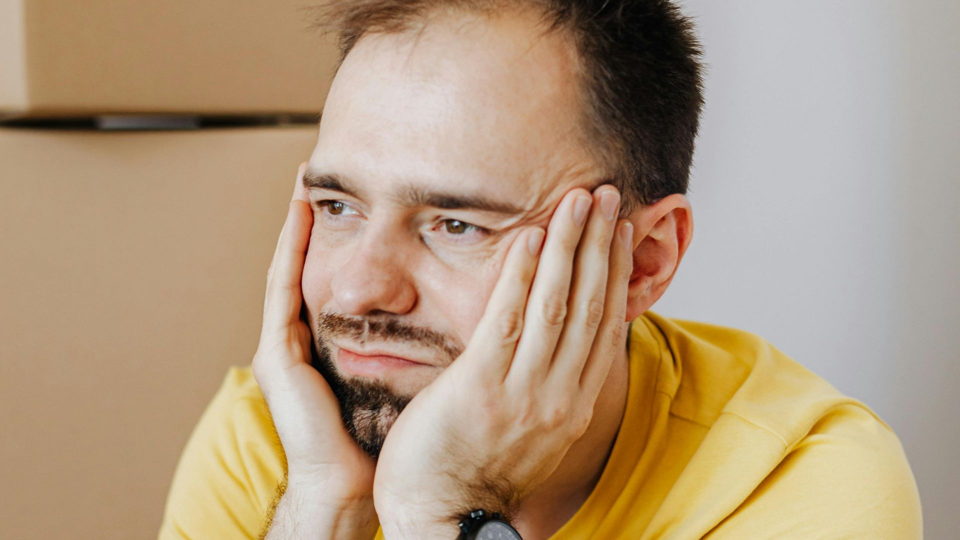 Frustrated man in casual wear holding face between hands while leaning on carton box and looking away with sadness after long tiring relocation day