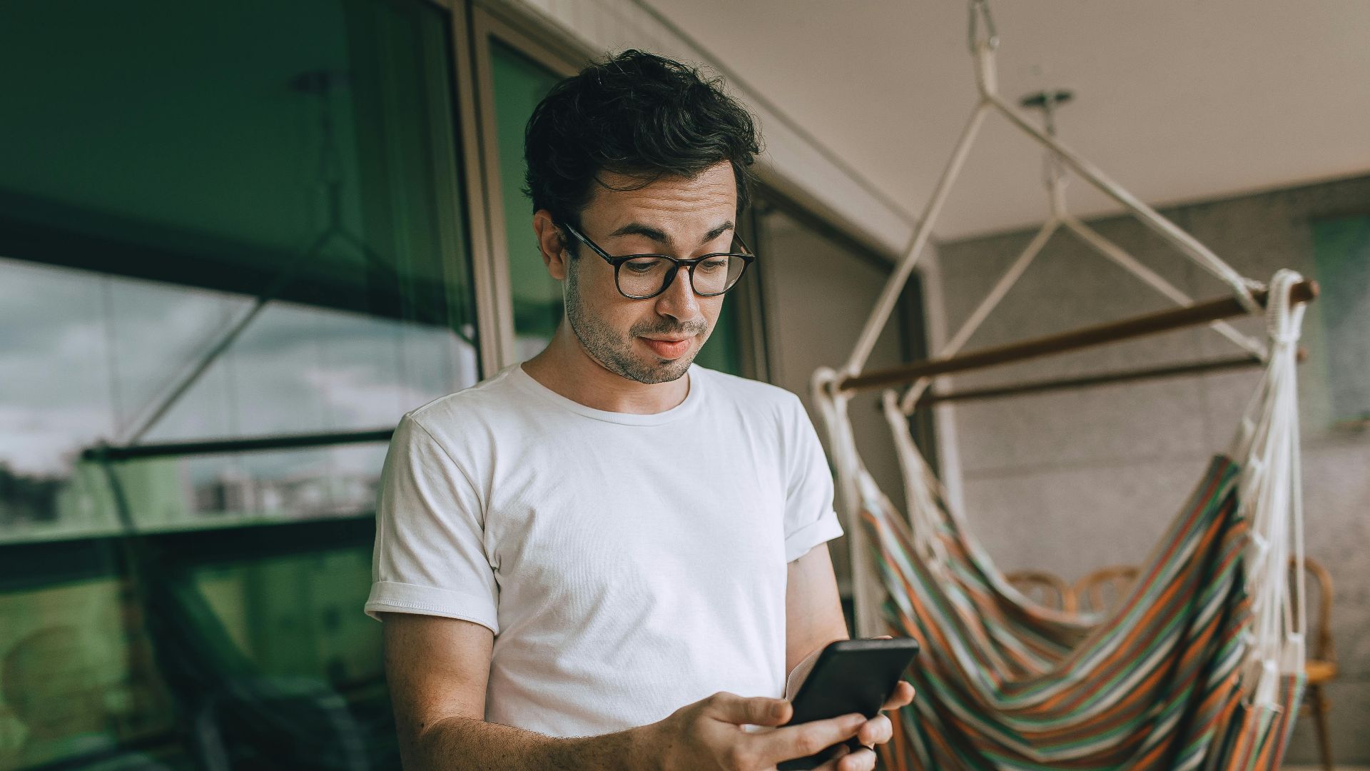 Young man in glasses using smartphone indoors with striped hammock.