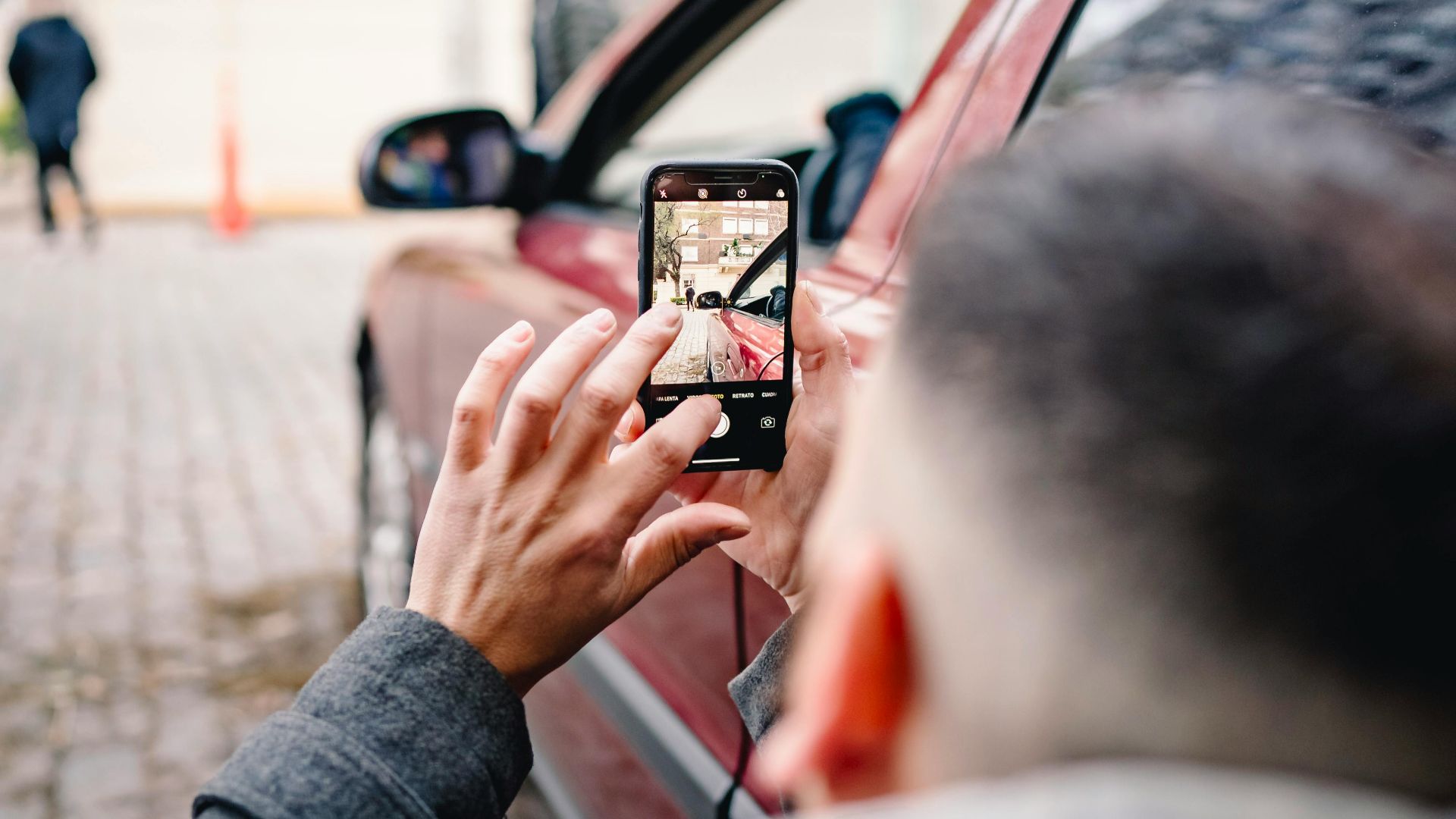 Man using smartphone to take pictures of an urban scene beside a red car.