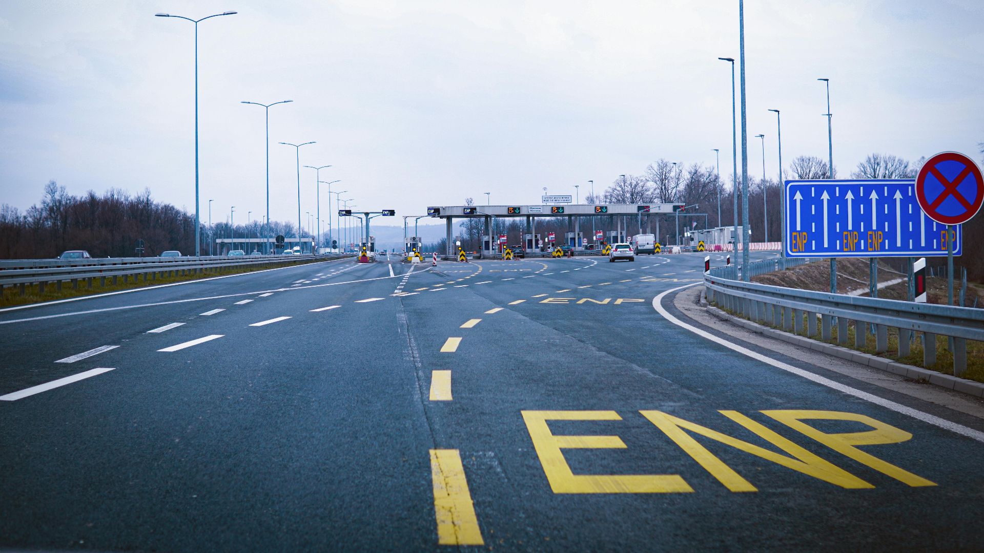 Wide highway leading to a toll plaza under a cloudy sky, featuring road signs and empty lanes.