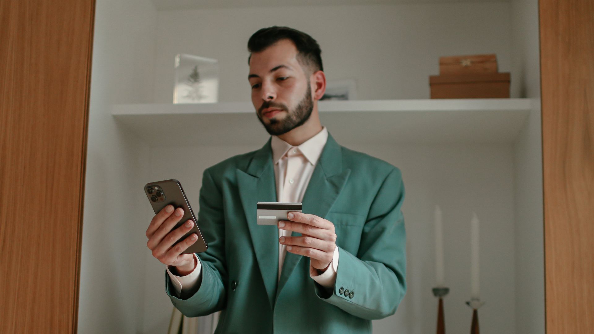 A bearded man in a green suit is using a smartphone and credit card indoors, showcasing modern technology and lifestyle.
