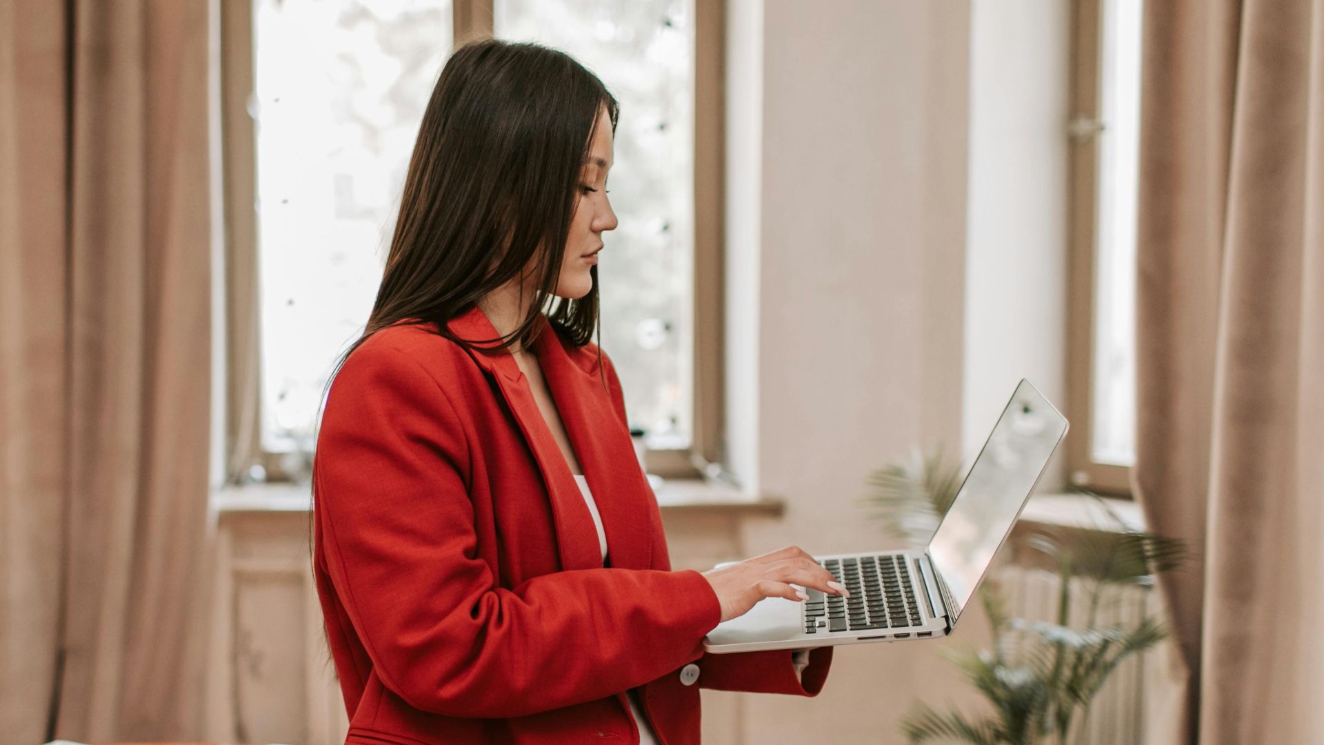 Woman in red blazer using laptop in modern office setting, multitasking efficiently.