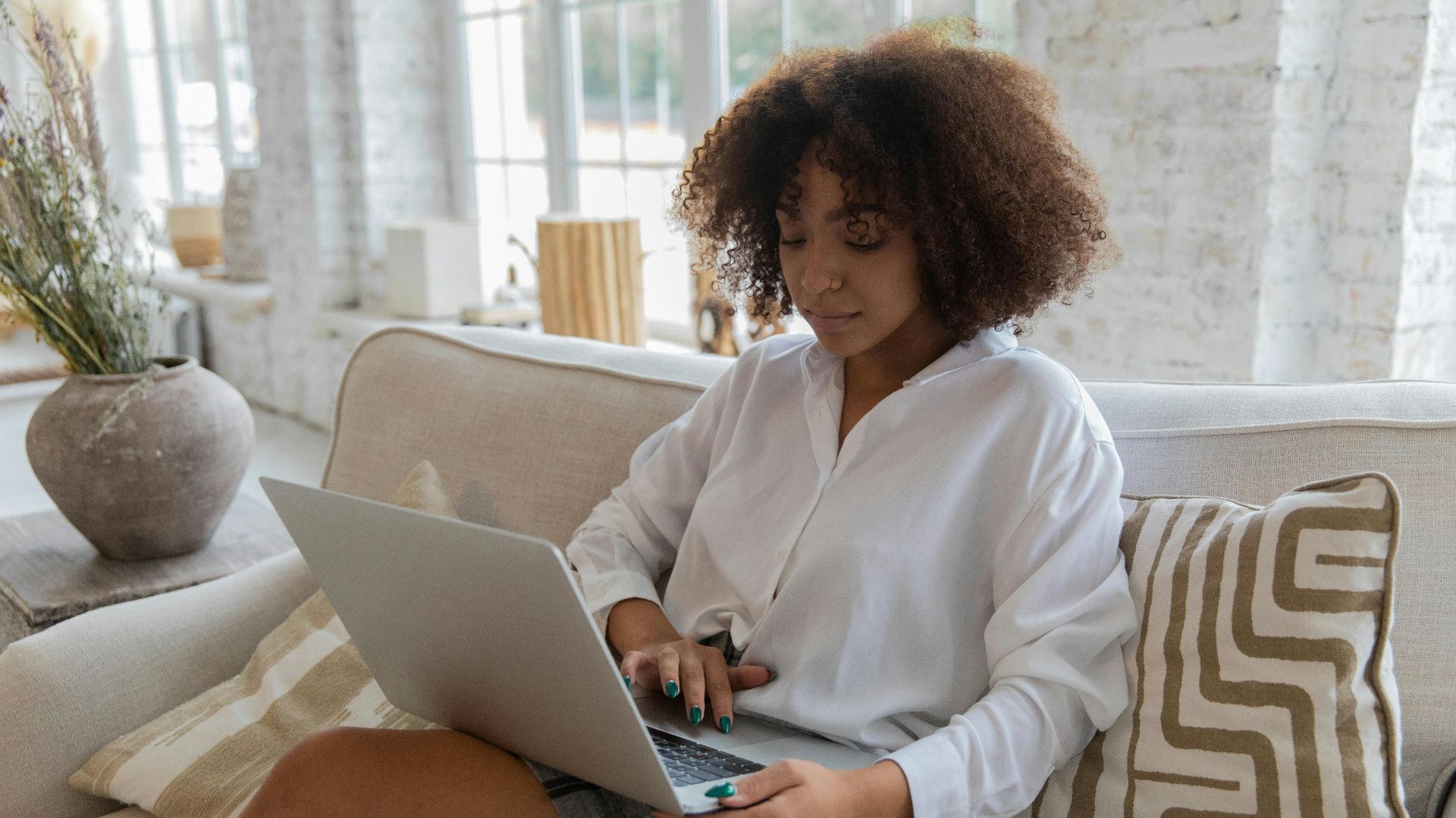Concentrated young African American female student with curly hair in casual clothes browsing laptop while sitting on sofa during online lesson at home