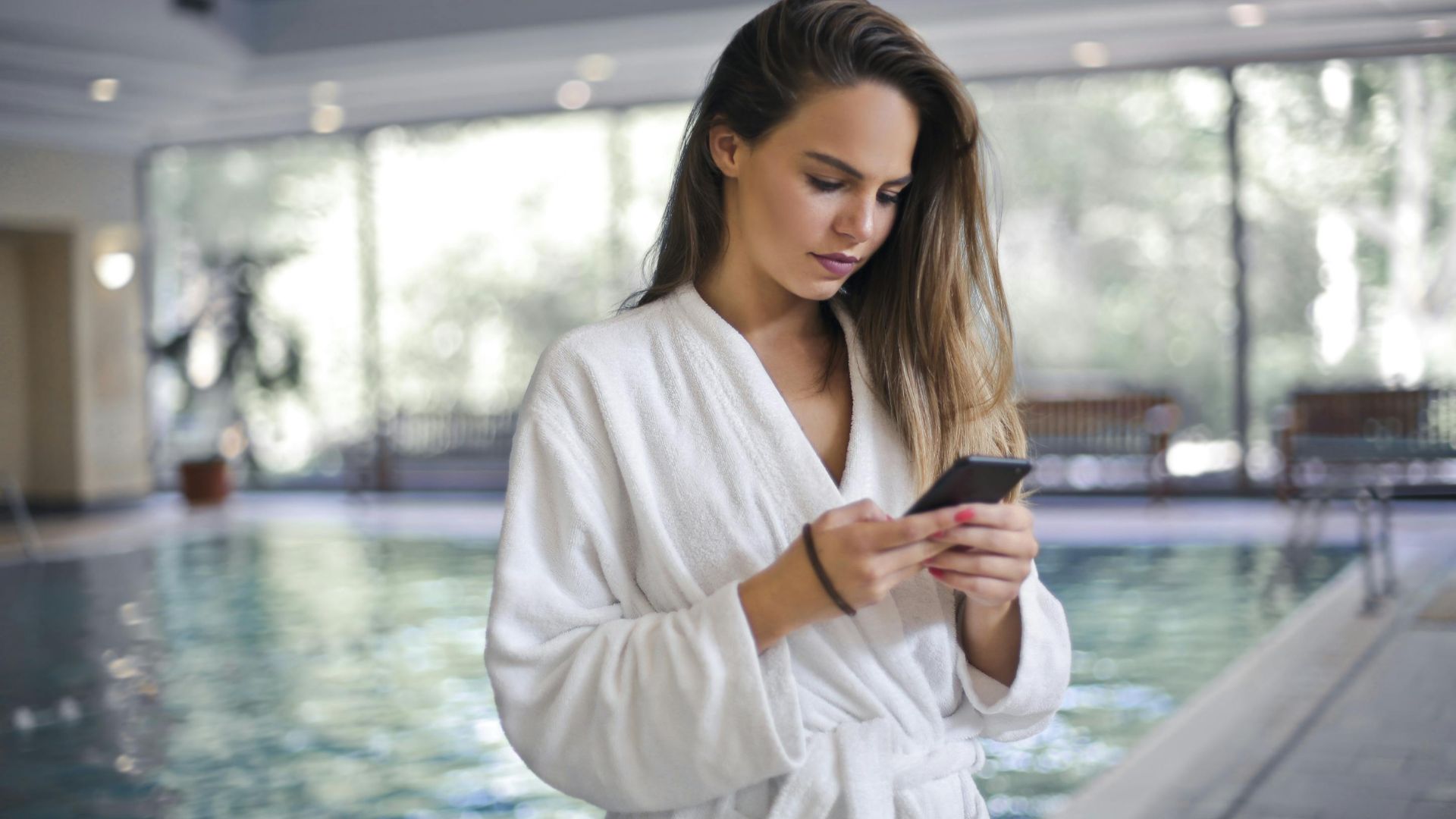 Woman in bathrobe by pool, texting on smartphone indoors. Calm and modern setting.
