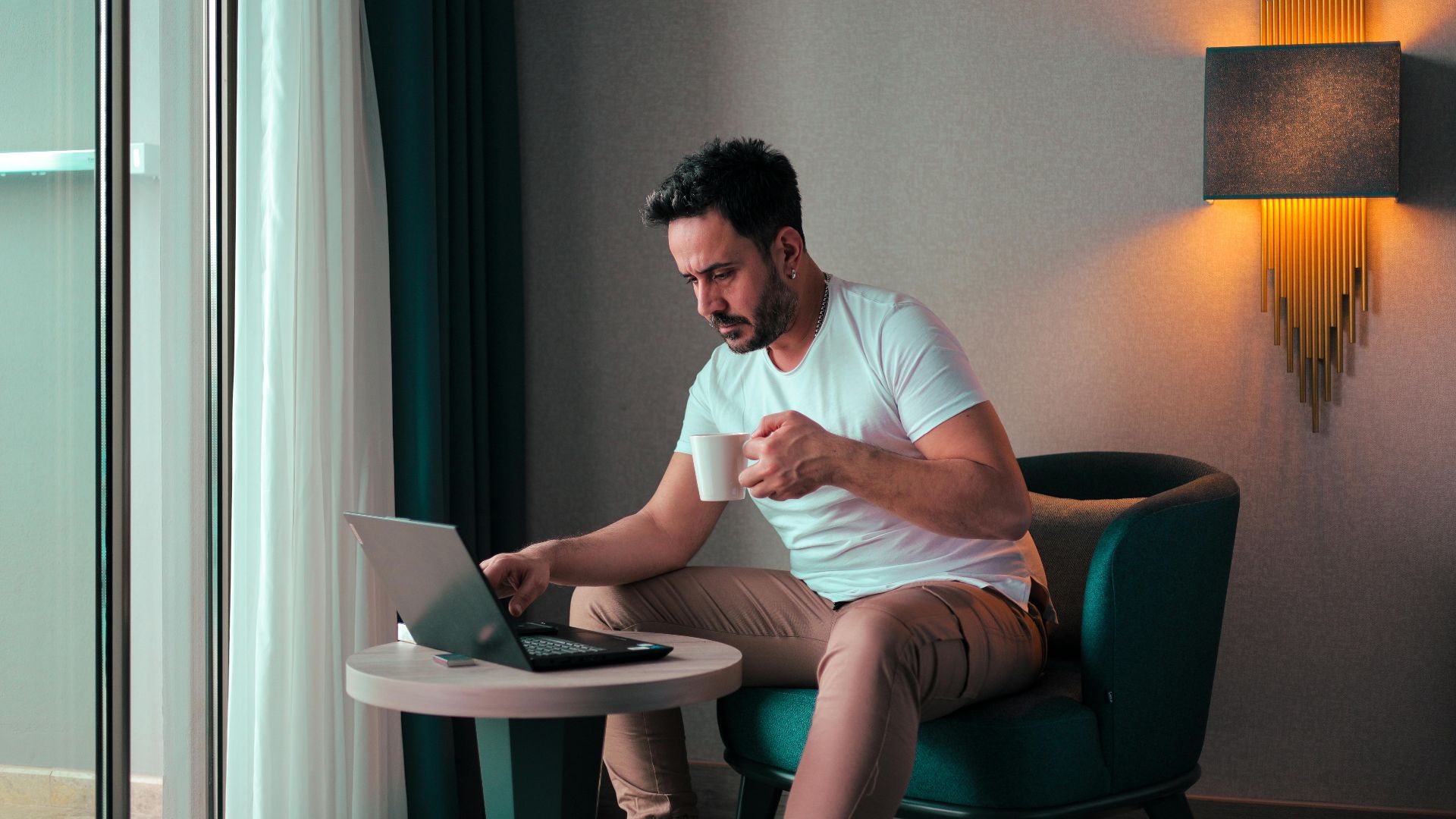 A man sits in a cozy hotel room in Cyprus, working on a laptop while enjoying a coffee.