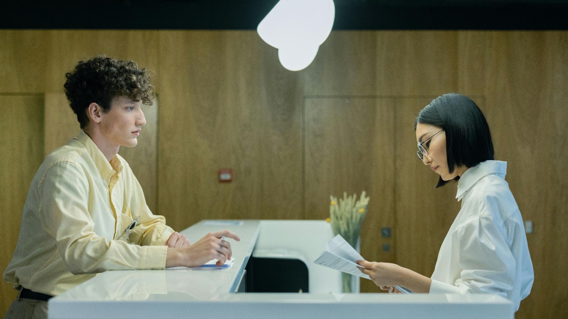 A young man and woman interacting at a modern office reception desk indoors.