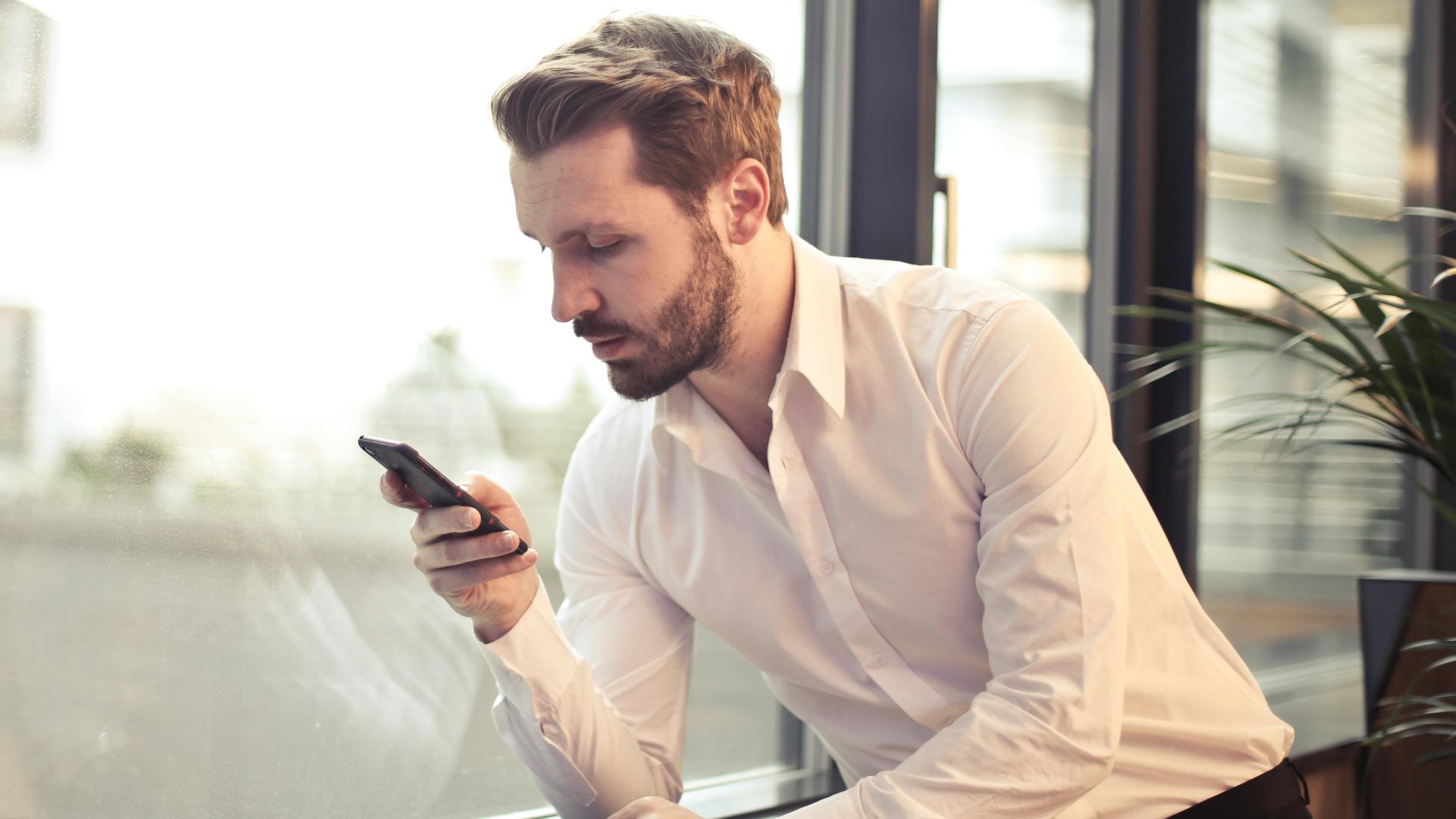 A businessman in a white shirt texts on his smartphone by a large glass window inside an office.