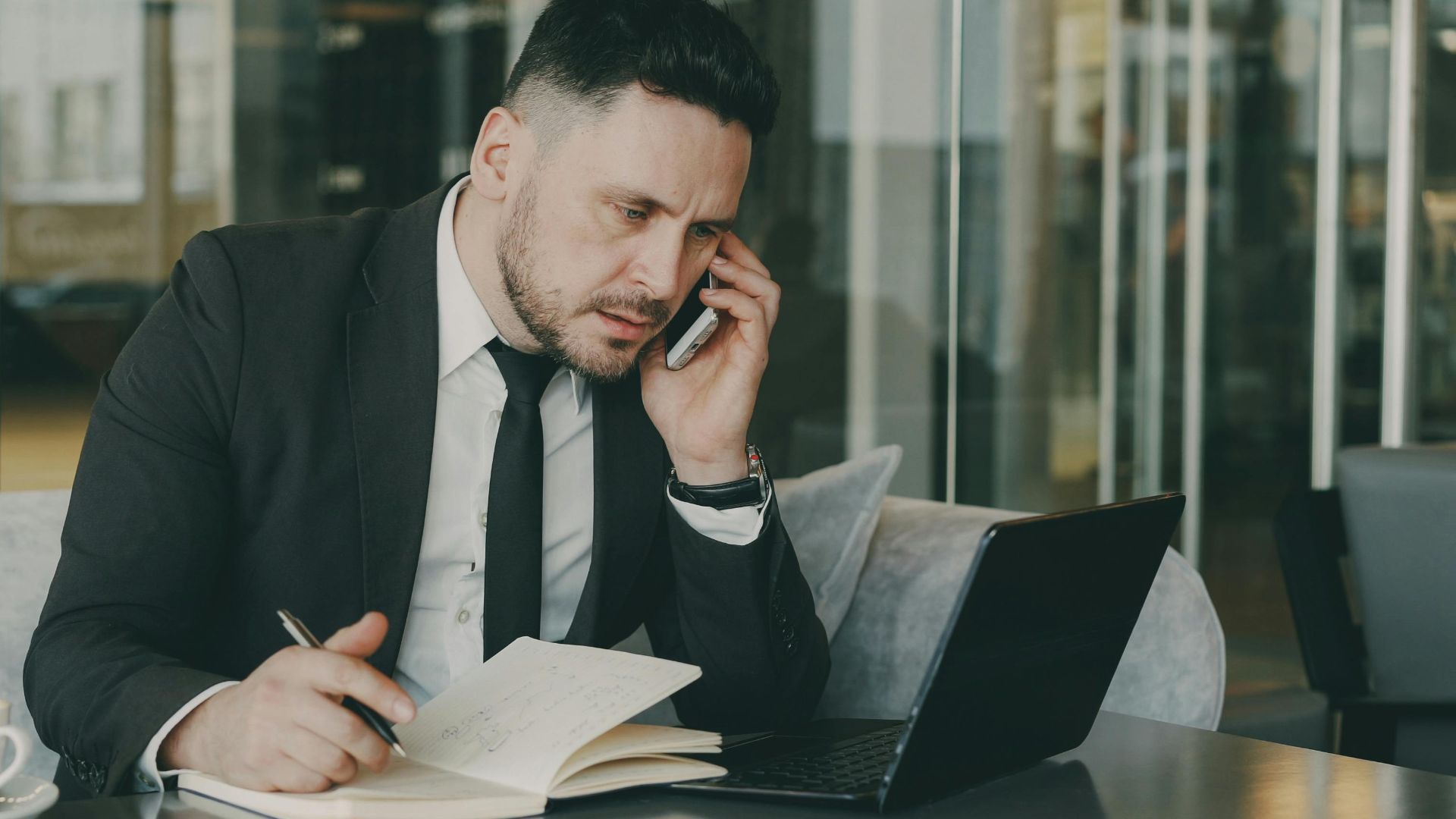 Businessman talks on phone while writing notes in a modern cafe.