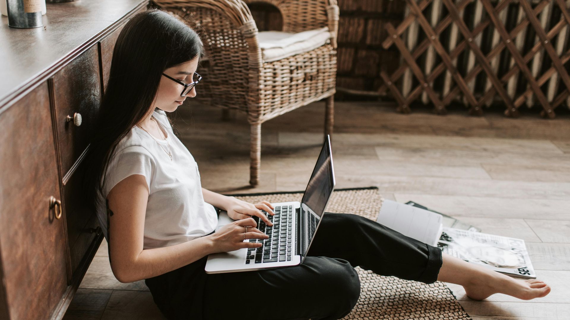 Side view of happy young woman with long dark hair in eyeglasses typing on keyboard of computer while sitting on floor in modern living room