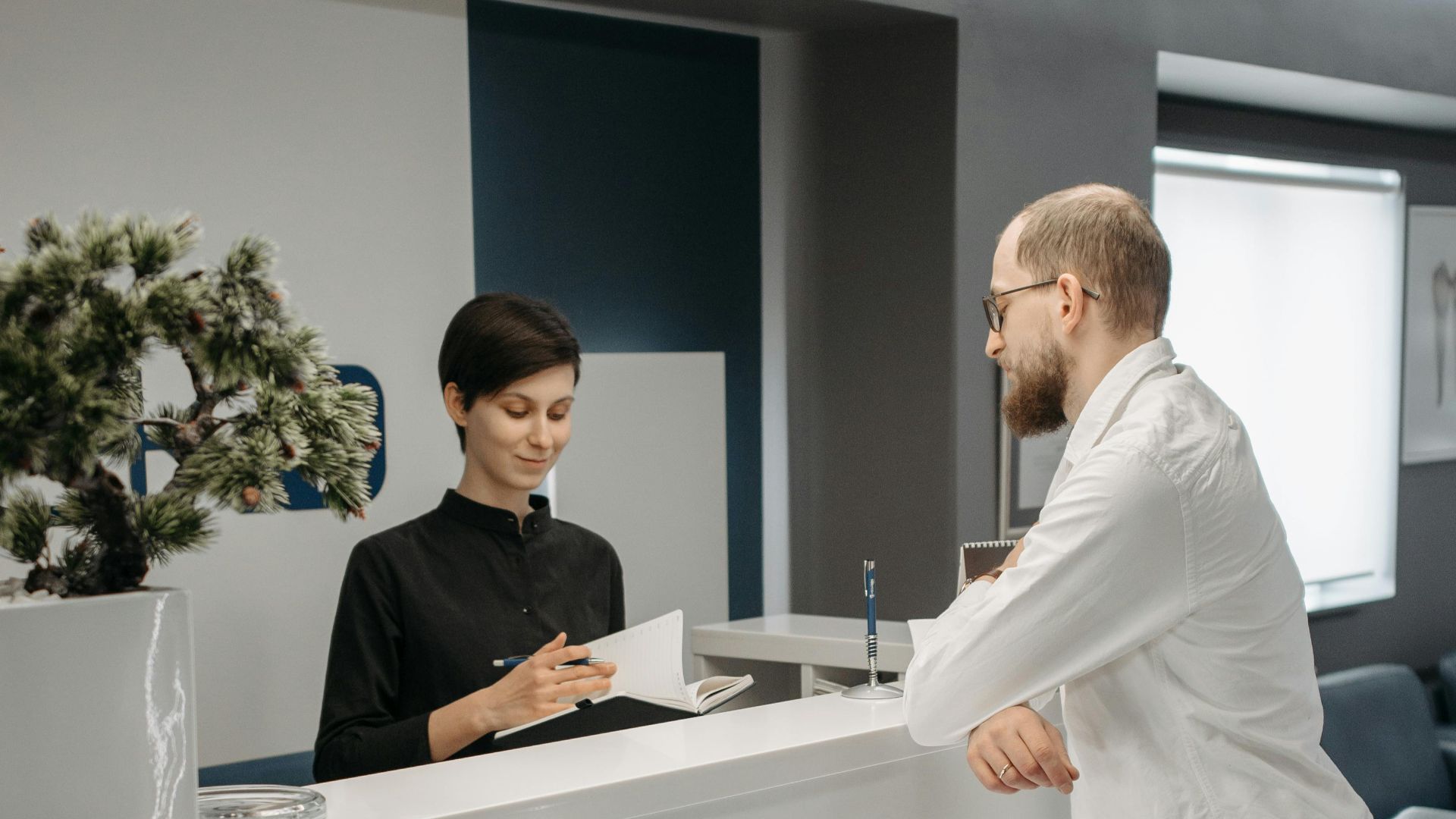 A man and woman at a sleek office reception desk, discussing paperwork in a professional setting.