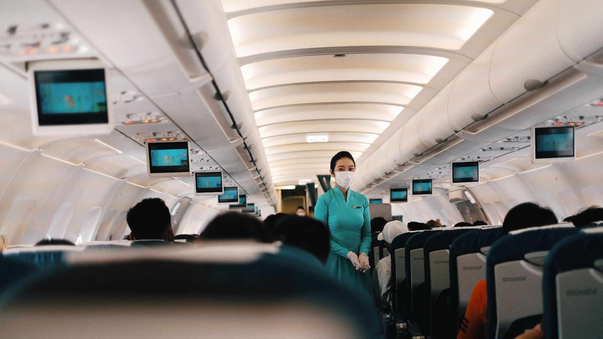 Flight attendant wearing a face mask attending to passengers in an airplane cabin.
