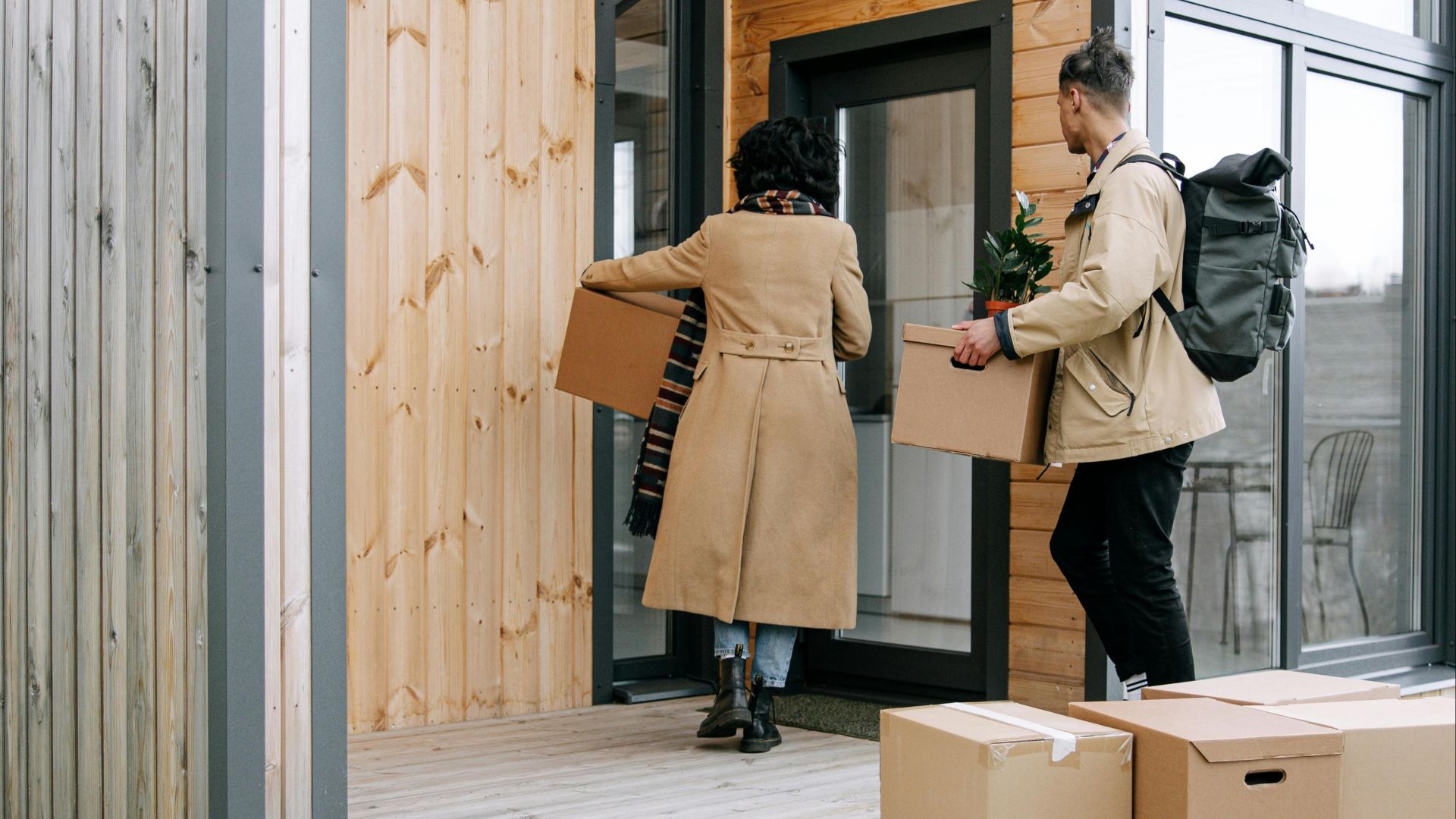 A couple carrying boxes and a plant into their new modern wooden home.