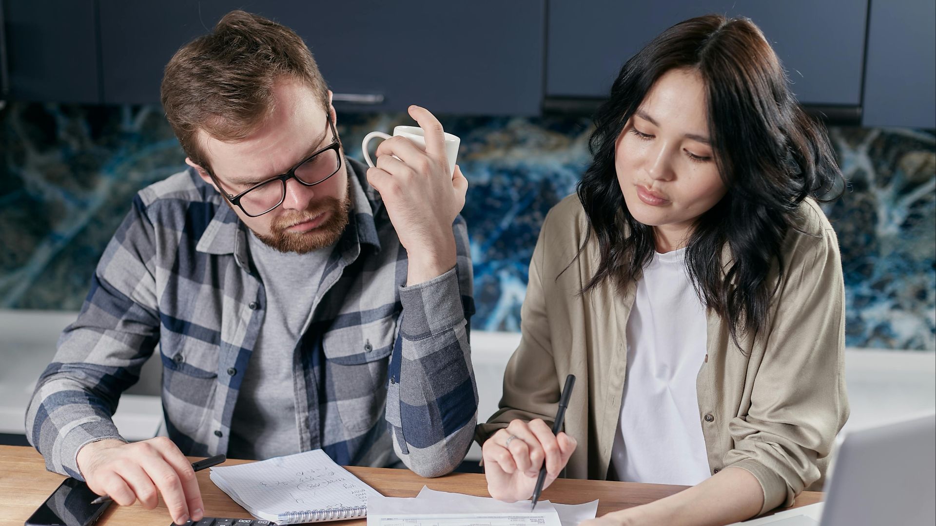 A couple reviewing household bills and budget using a calculator and laptop at their kitchen table.