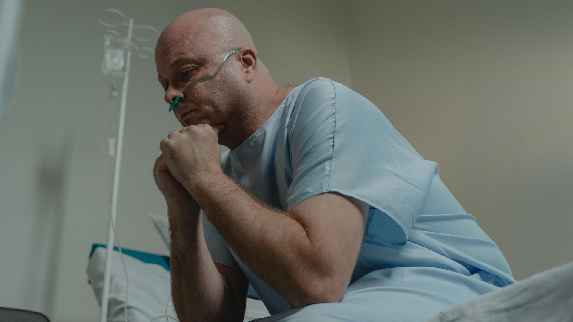 Bald man in hospital gown sits pensively with oxygen tube in healthcare setting.