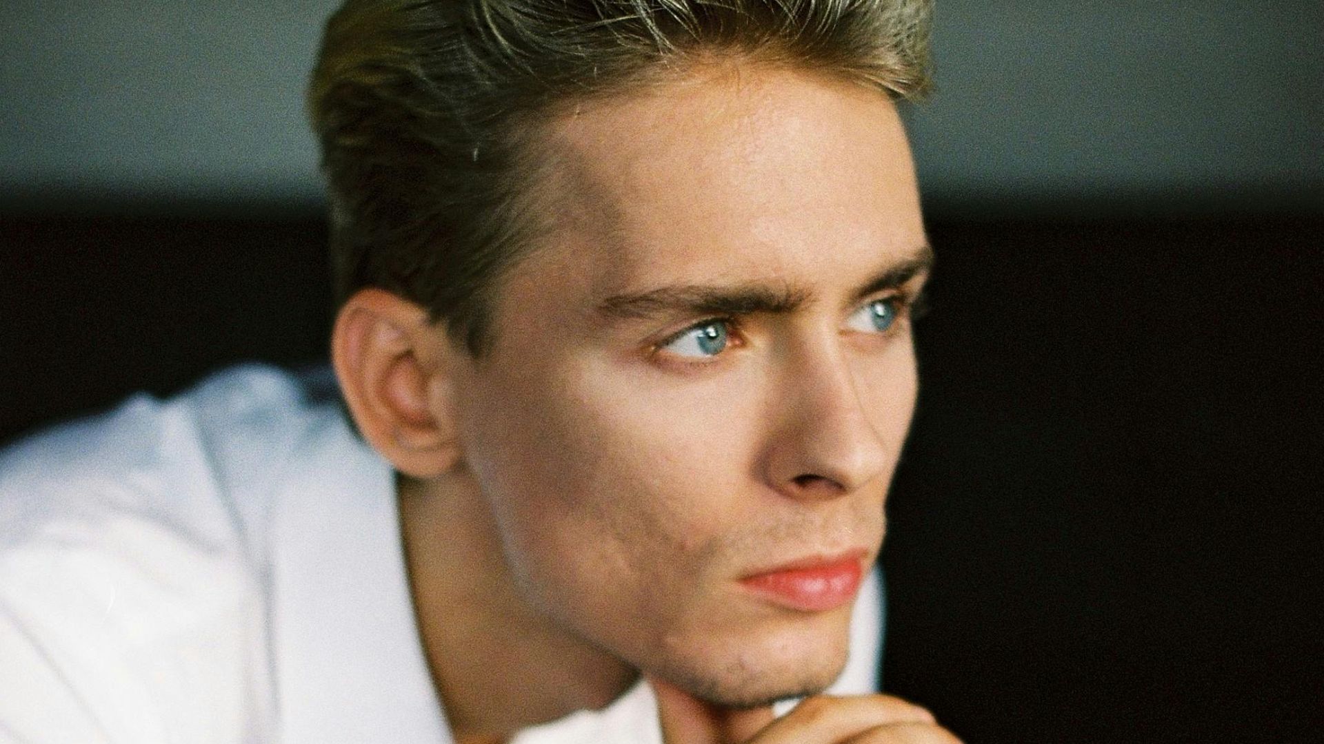 Handsome young guy with blond hair in elegant white shirt touching chin and looking away thoughtfully while sitting on chair in room