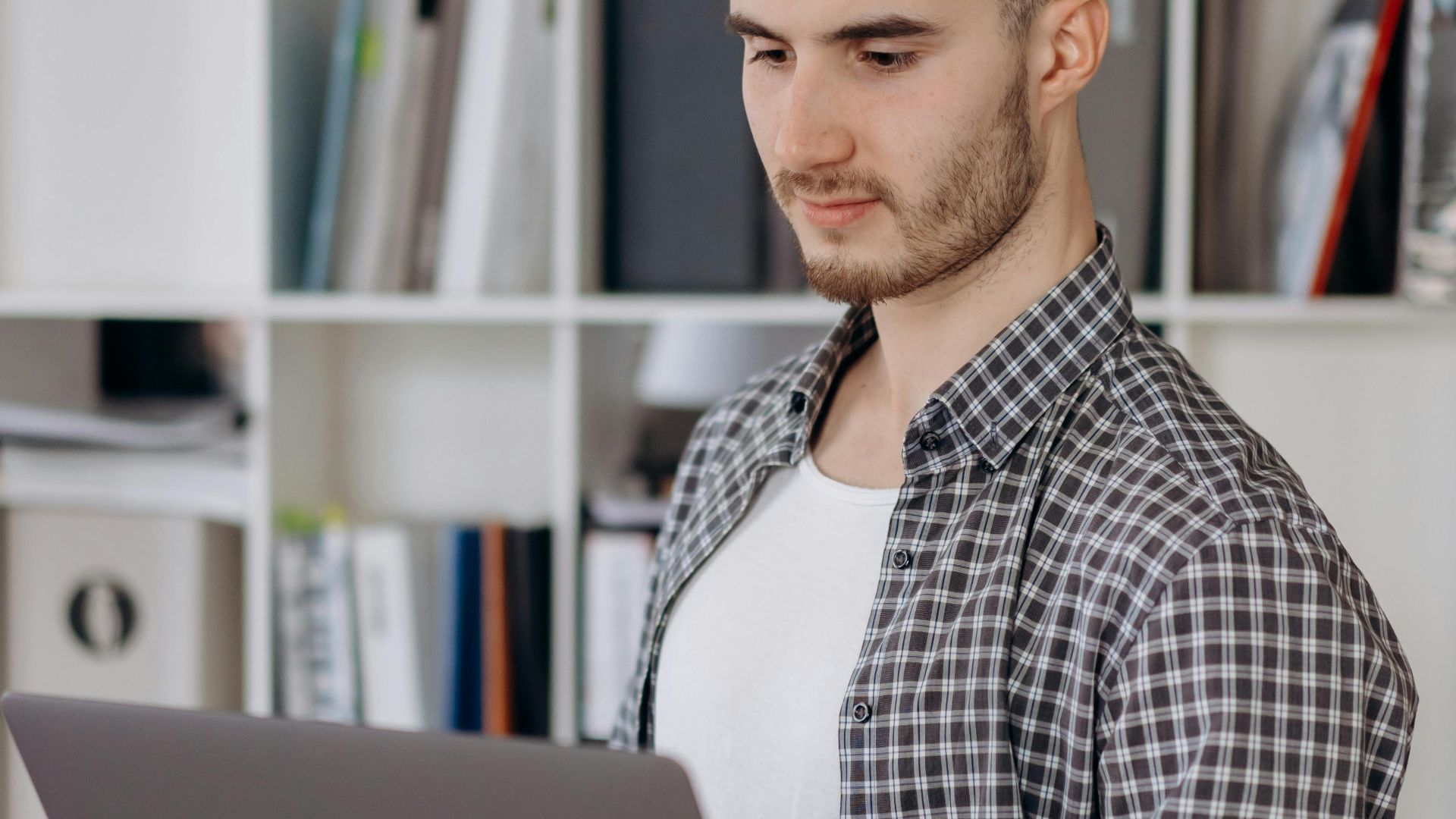 Young man attentively looking at laptop in a modern office with bookshelves.