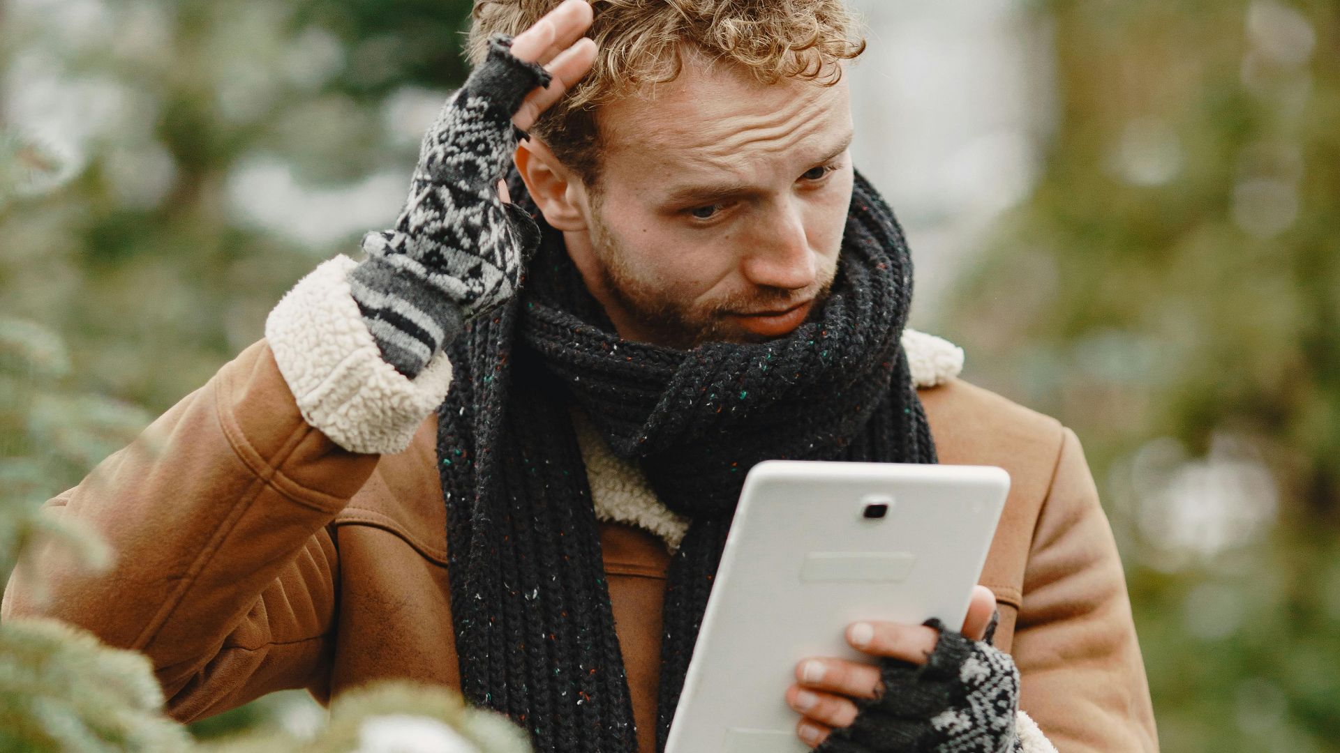 Man with a scarf using a tablet in a snowy forest, pondering or problem-solving.