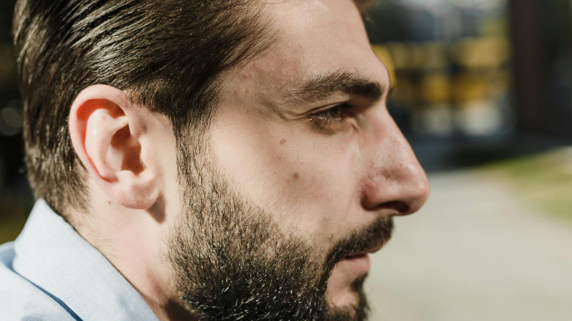Side profile of a bearded man in a light blue shirt outdoors, thoughtful expression.