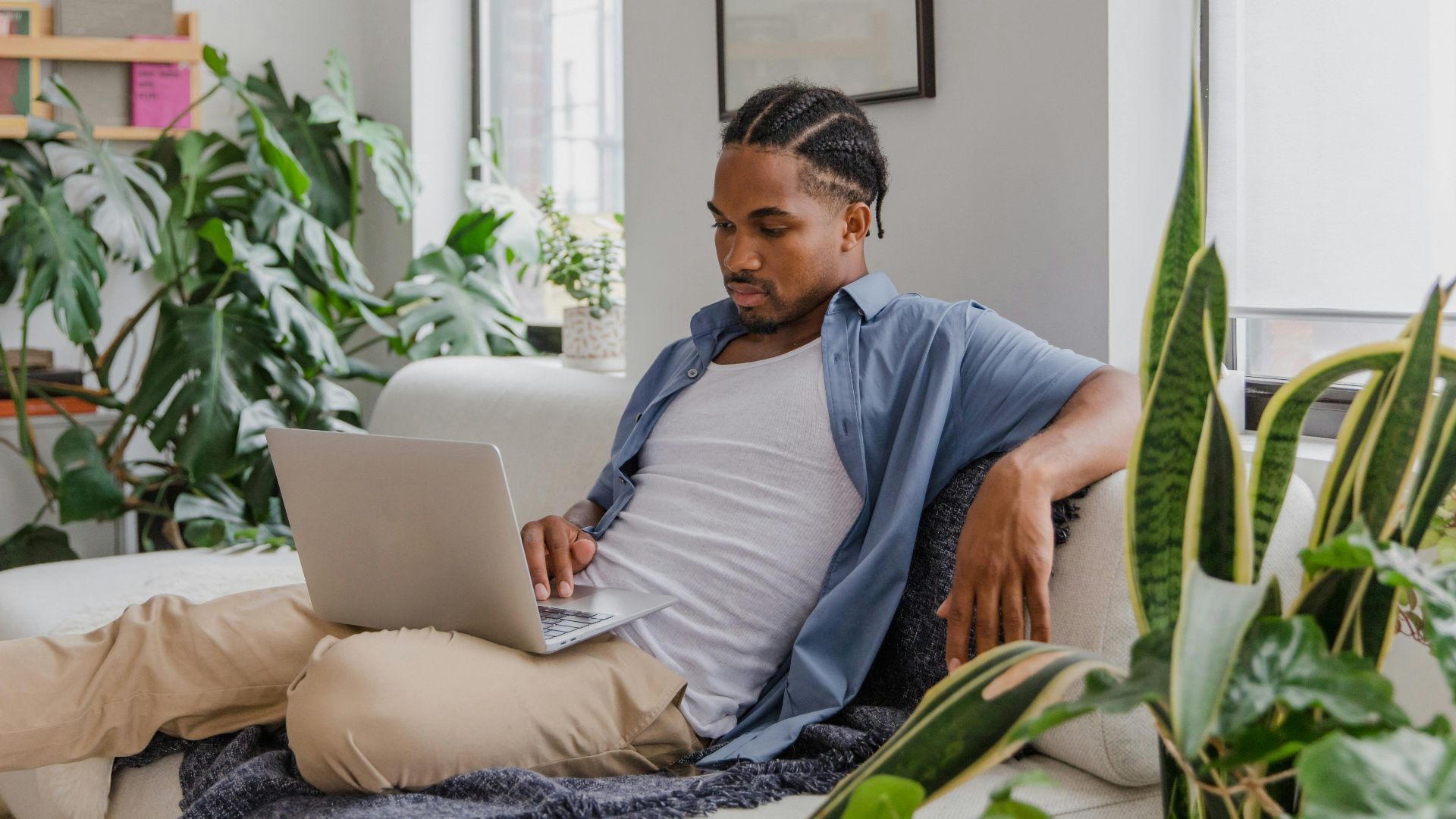 Casual man working from home office, surrounded by plants, on a comfortable sofa with a laptop.