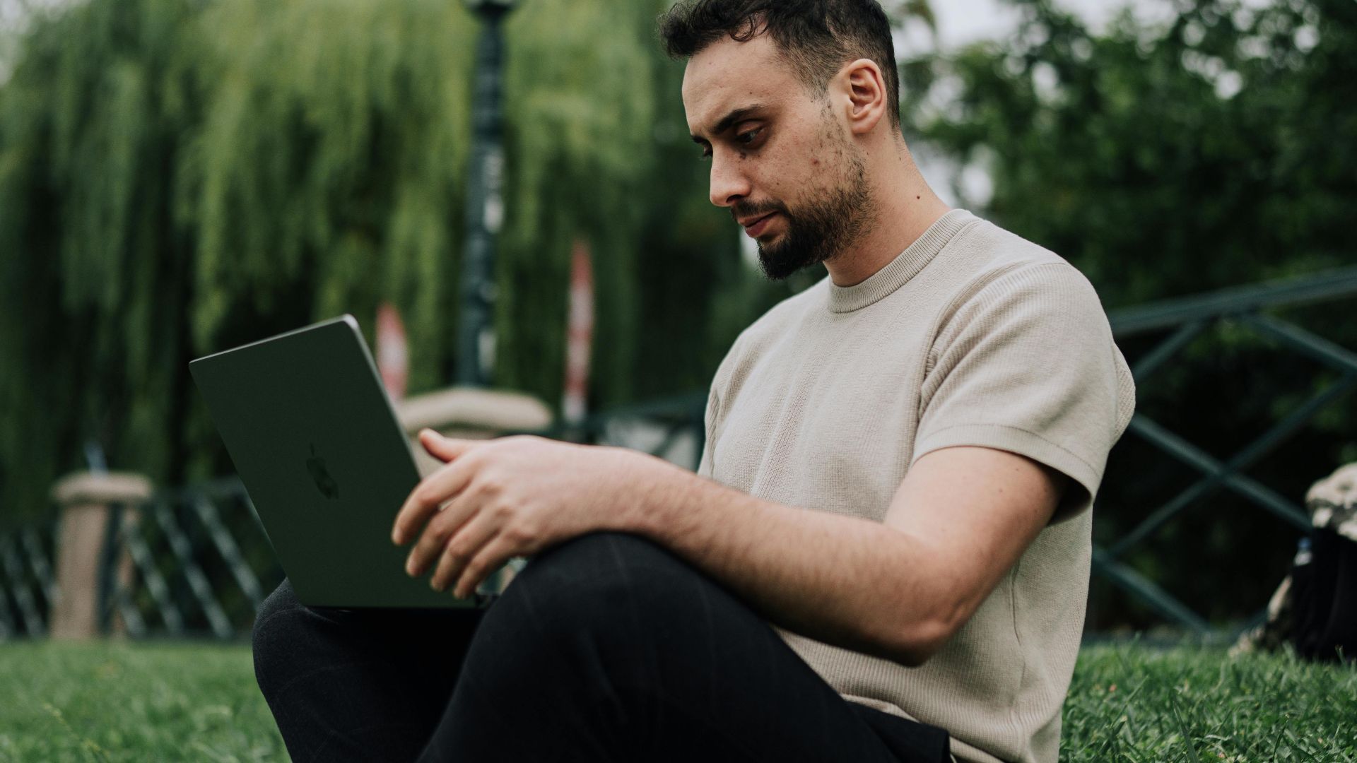 Casual man sitting on grass in park, browsing on laptop during a summer day.
