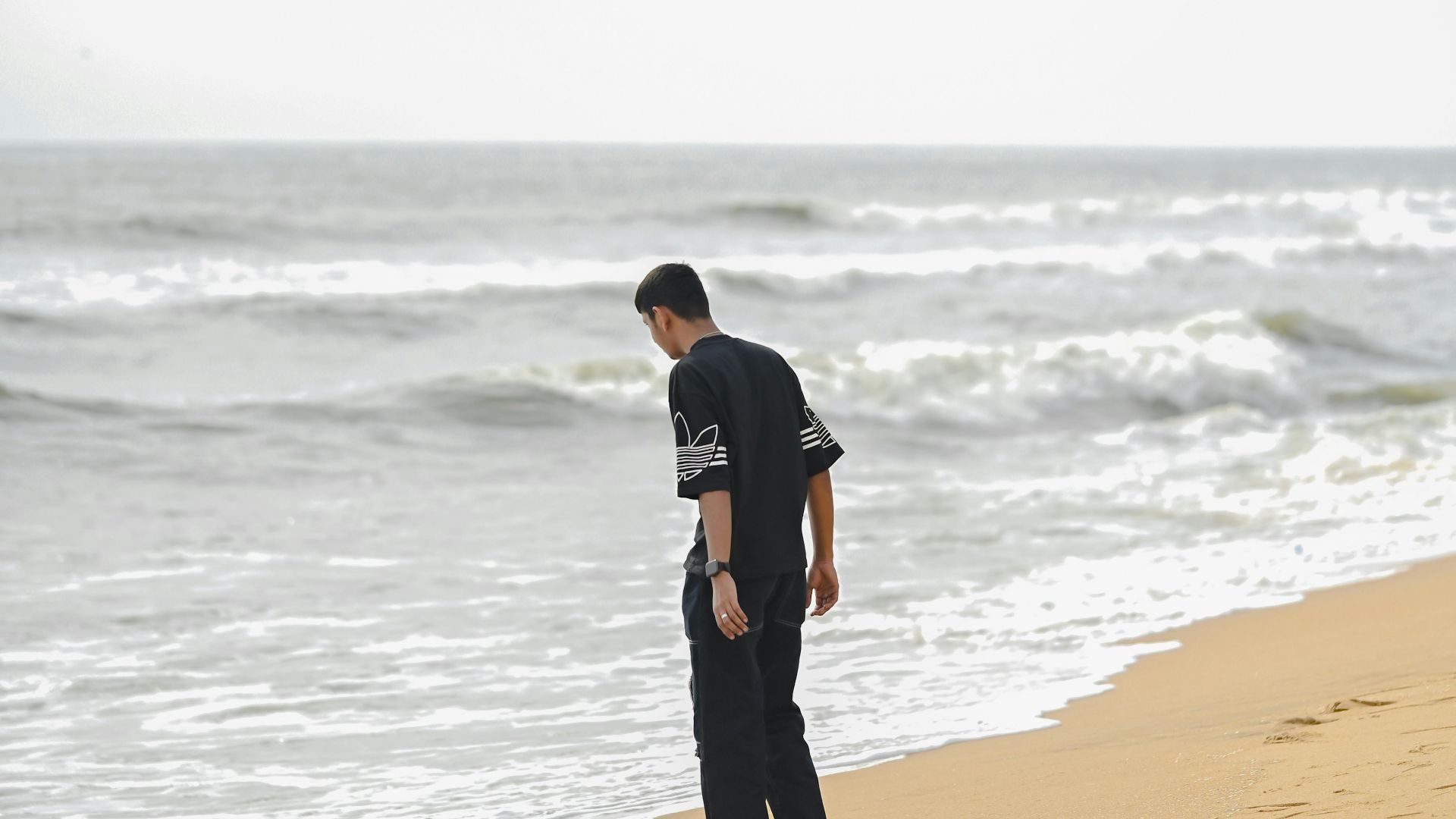 A solitary adult male walks along the beach, gazing at the ocean under a cloudy sky.
