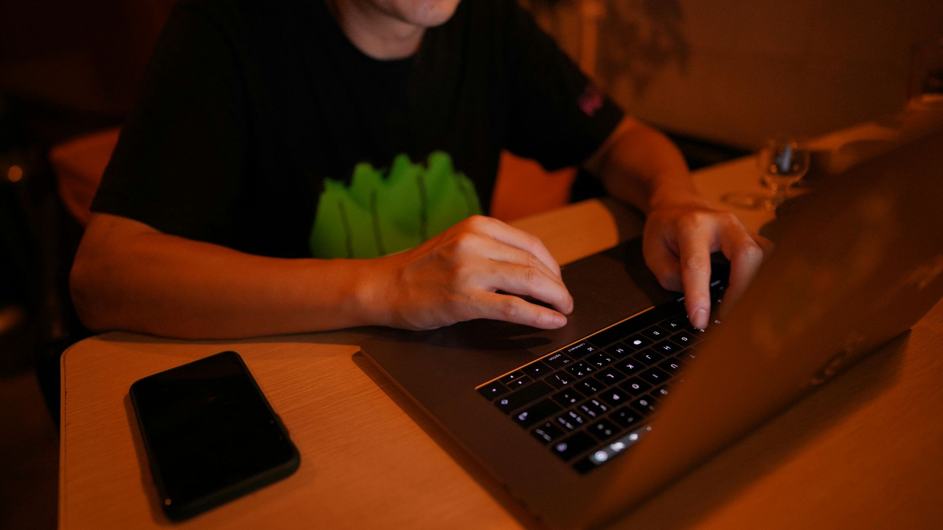 An adult man working at a desk on a laptop with a smartphone nearby, in a dimly lit home office.