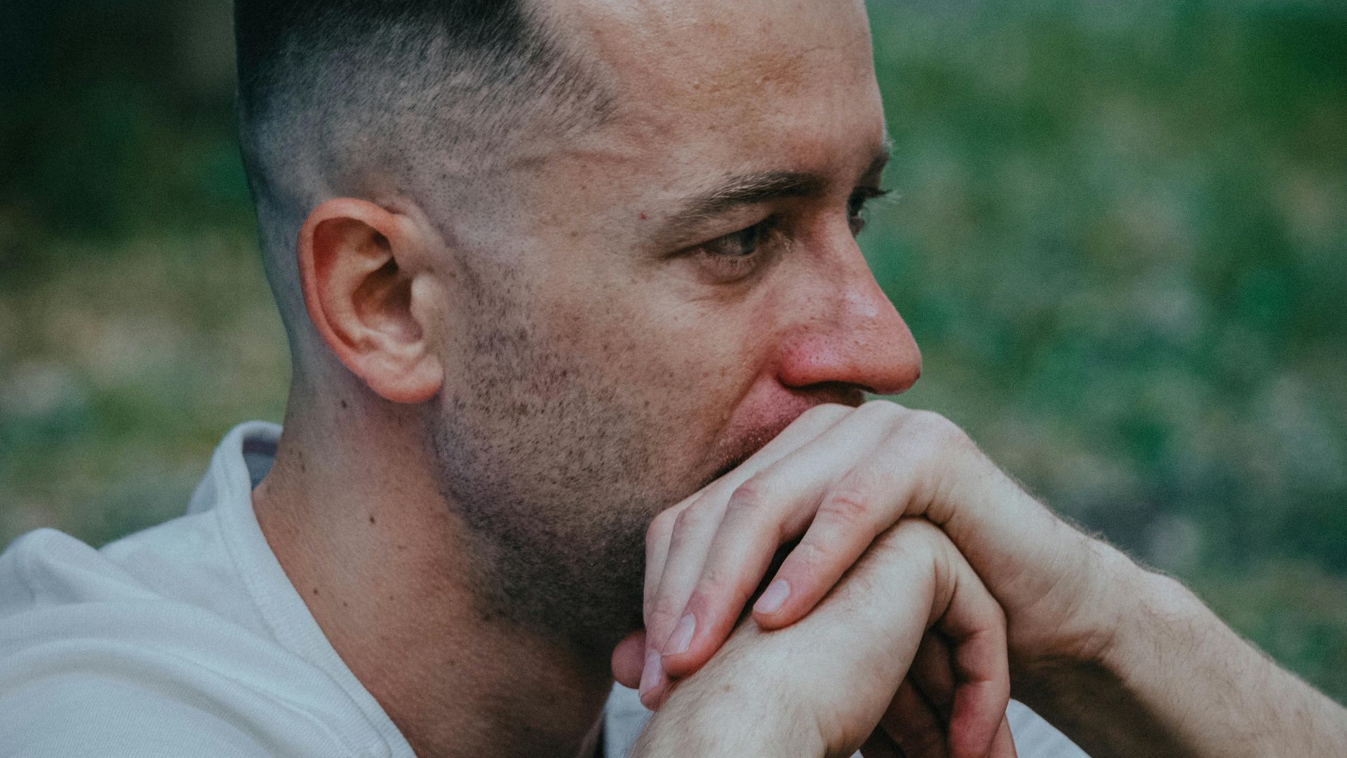 A thoughtful man outdoors, hands clasped near face, reflecting deeply.