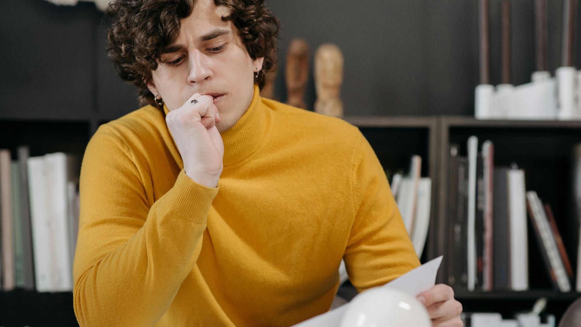 A thoughtful man in a yellow sweater closely examines a paper, seated indoors with books in the background.