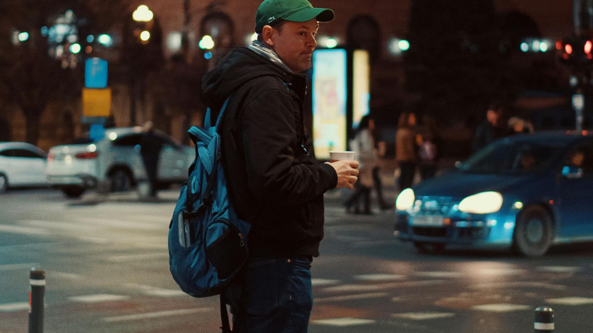 A man in a hoodie and backpack stands on a city street in the evening, holding a coffee.