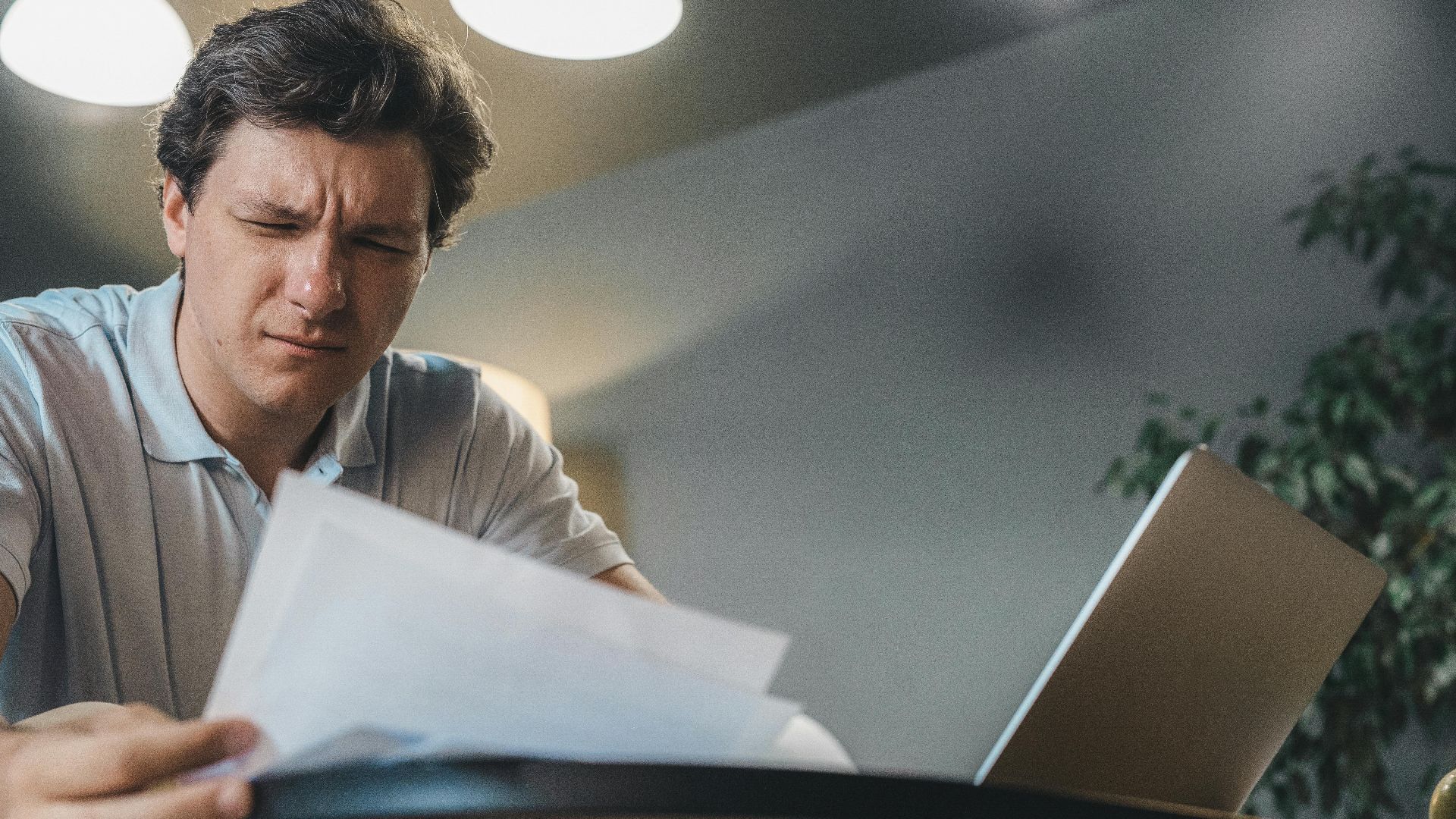 Man focusing intently while reviewing documents at a desk with a laptop.