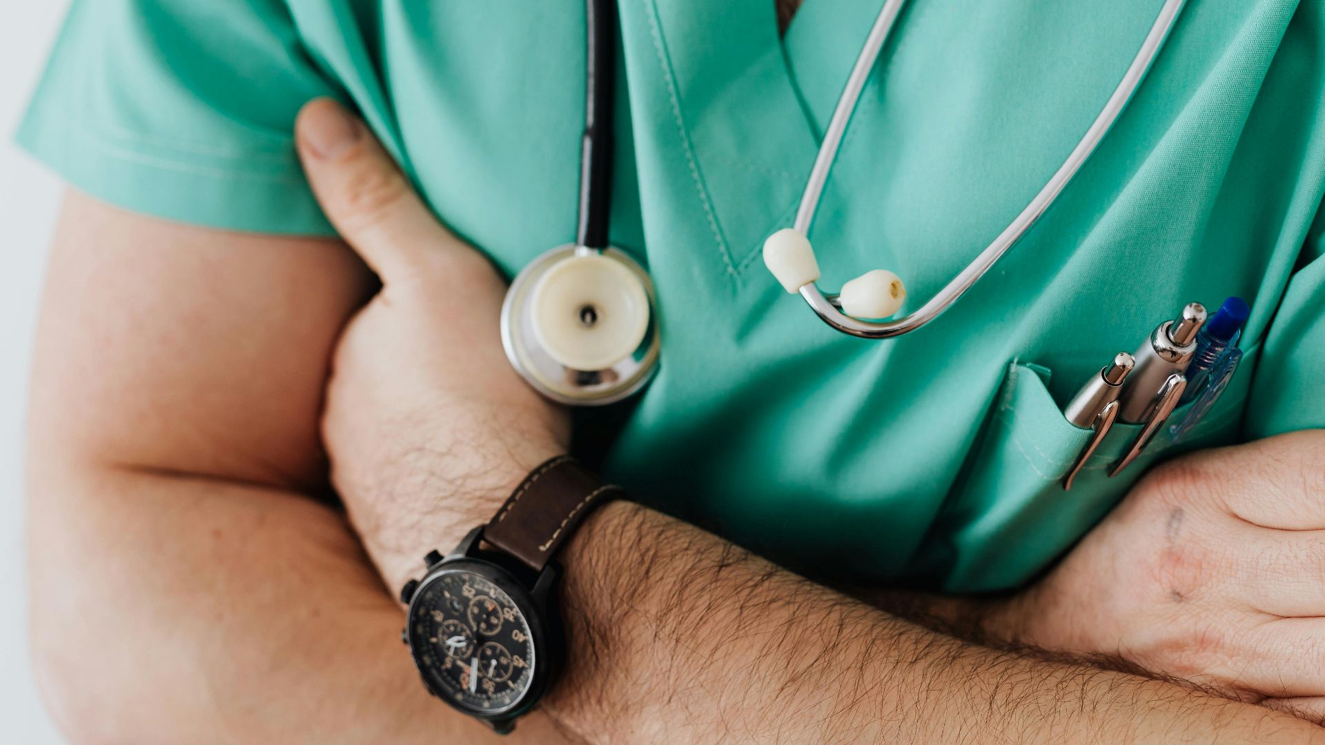 Unrecognizable man wearing medical uniform with stethoscope and pens standing in hospital with crossed arms during work in day time