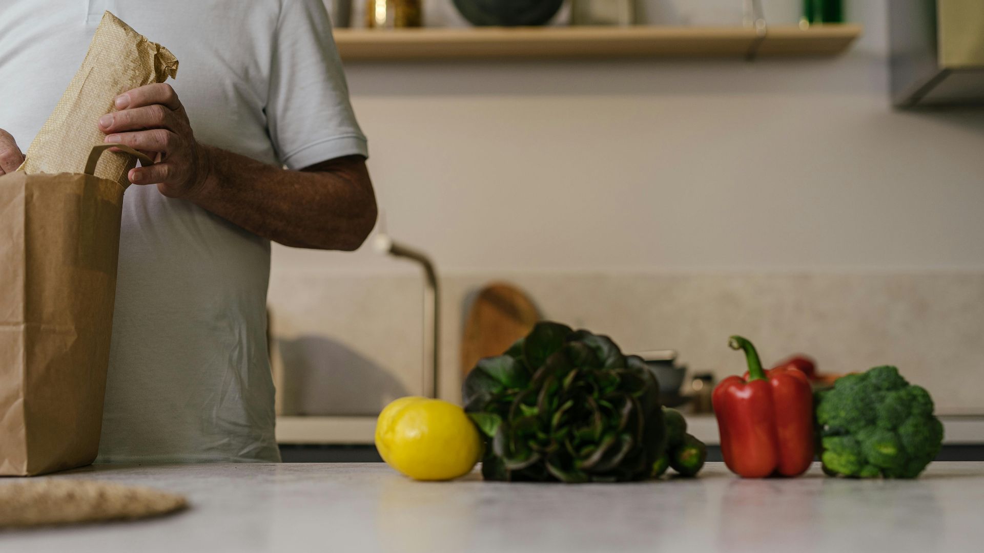 A man unpacks groceries from a paper bag on a kitchen counter with fresh vegetables.