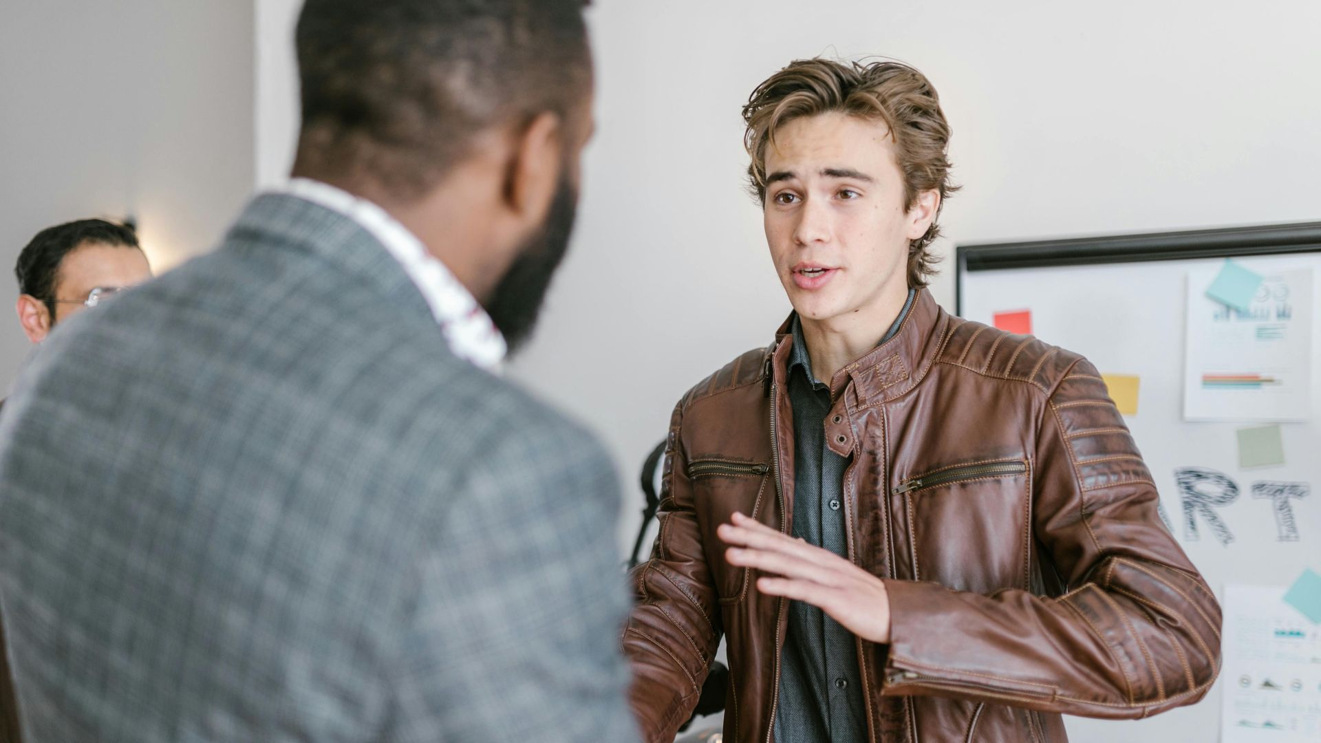 A young man in a leather jacket talking with colleagues during a business meeting indoors.