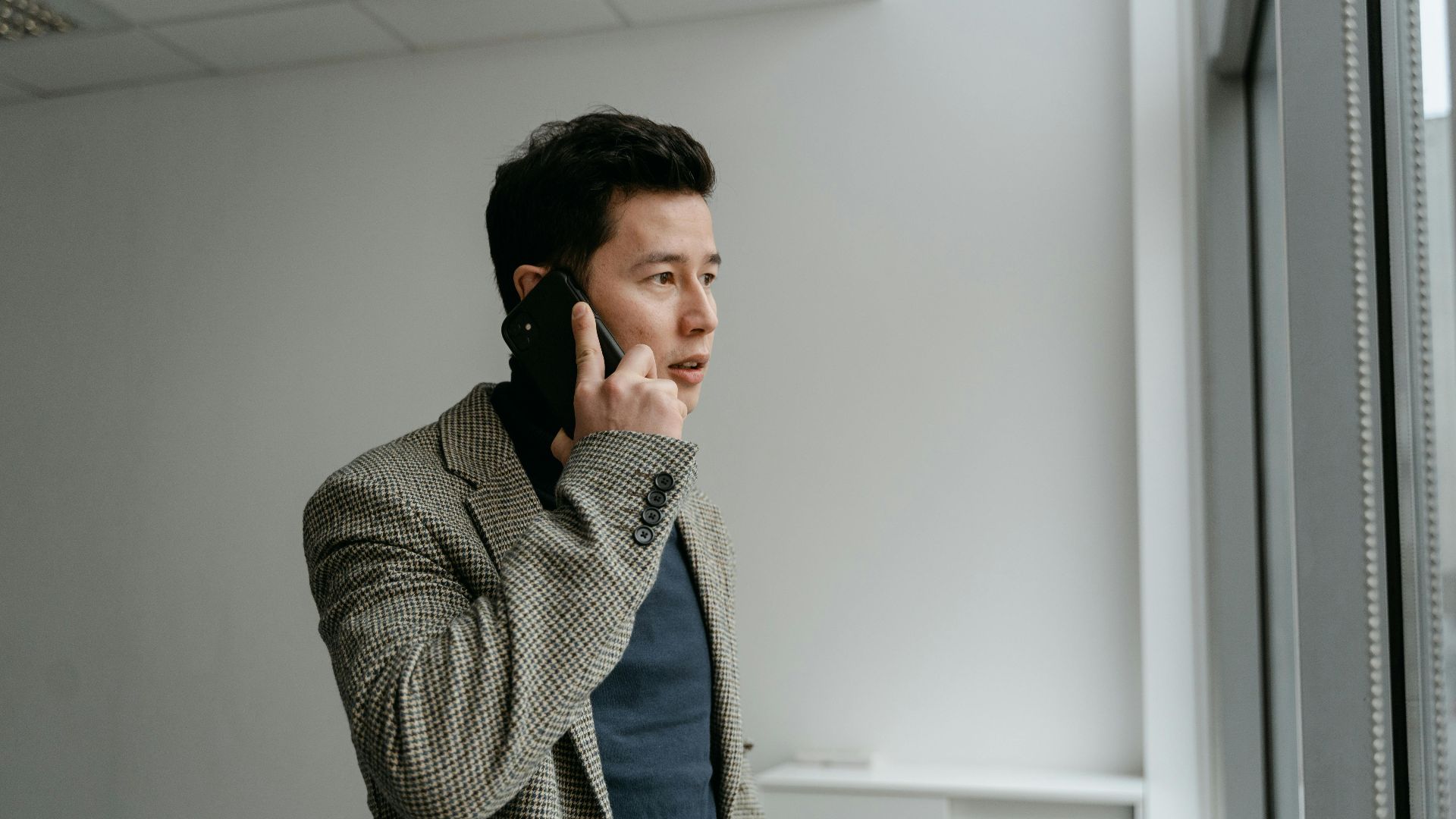 Young businessman in blazer talking on smartphone near window indoors.