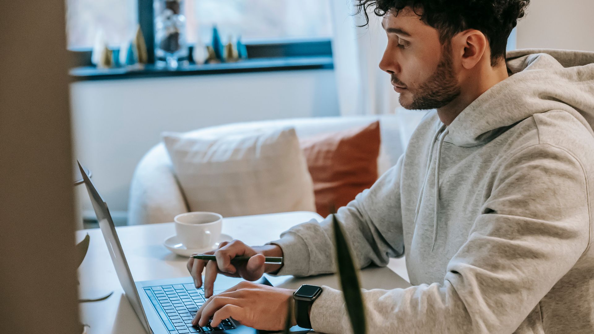 Side view of crop focused male remote worker browsing internet on netbook at table with coffee in house