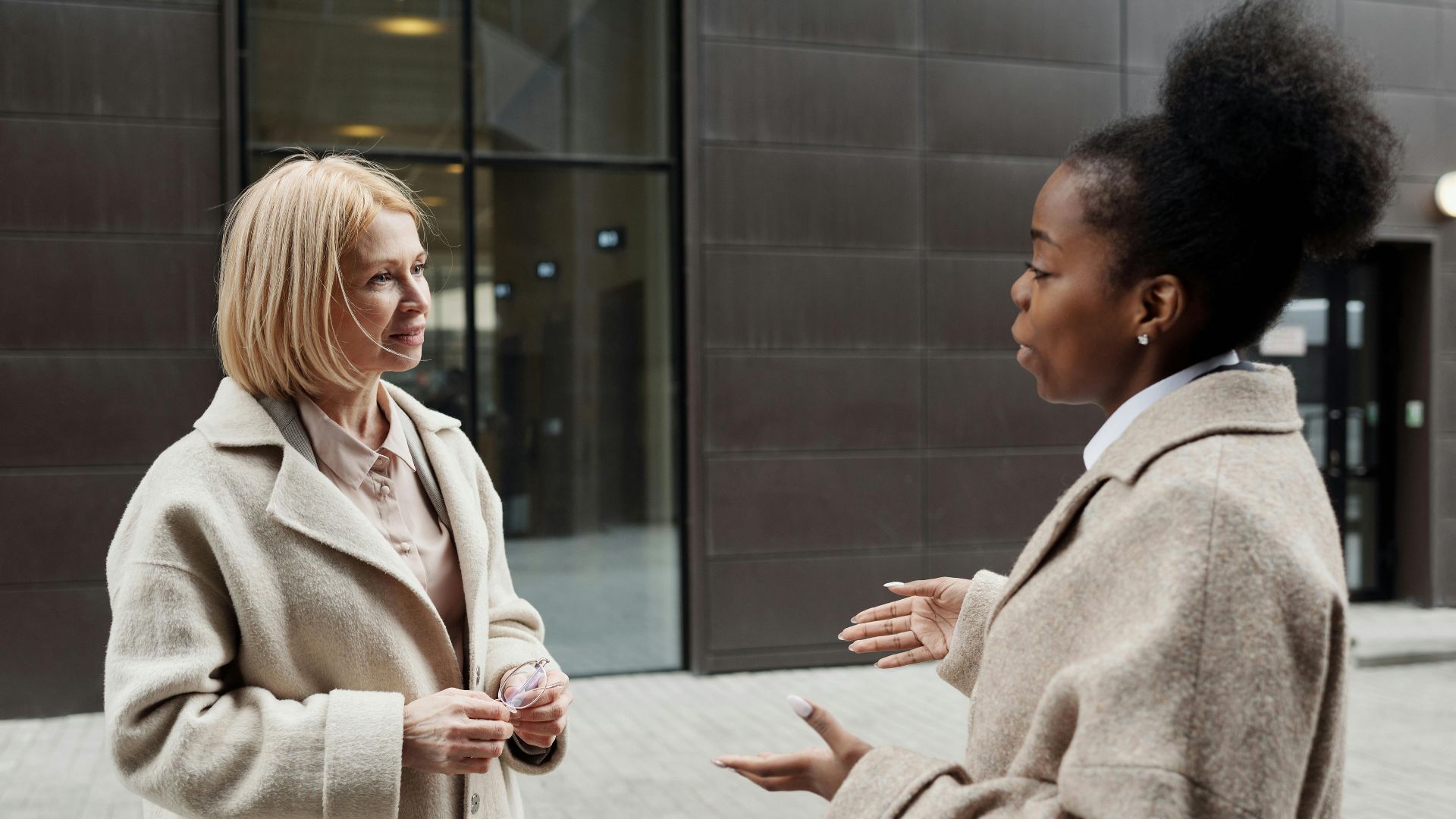 Two businesswomen in coats converse outside a modern office building.