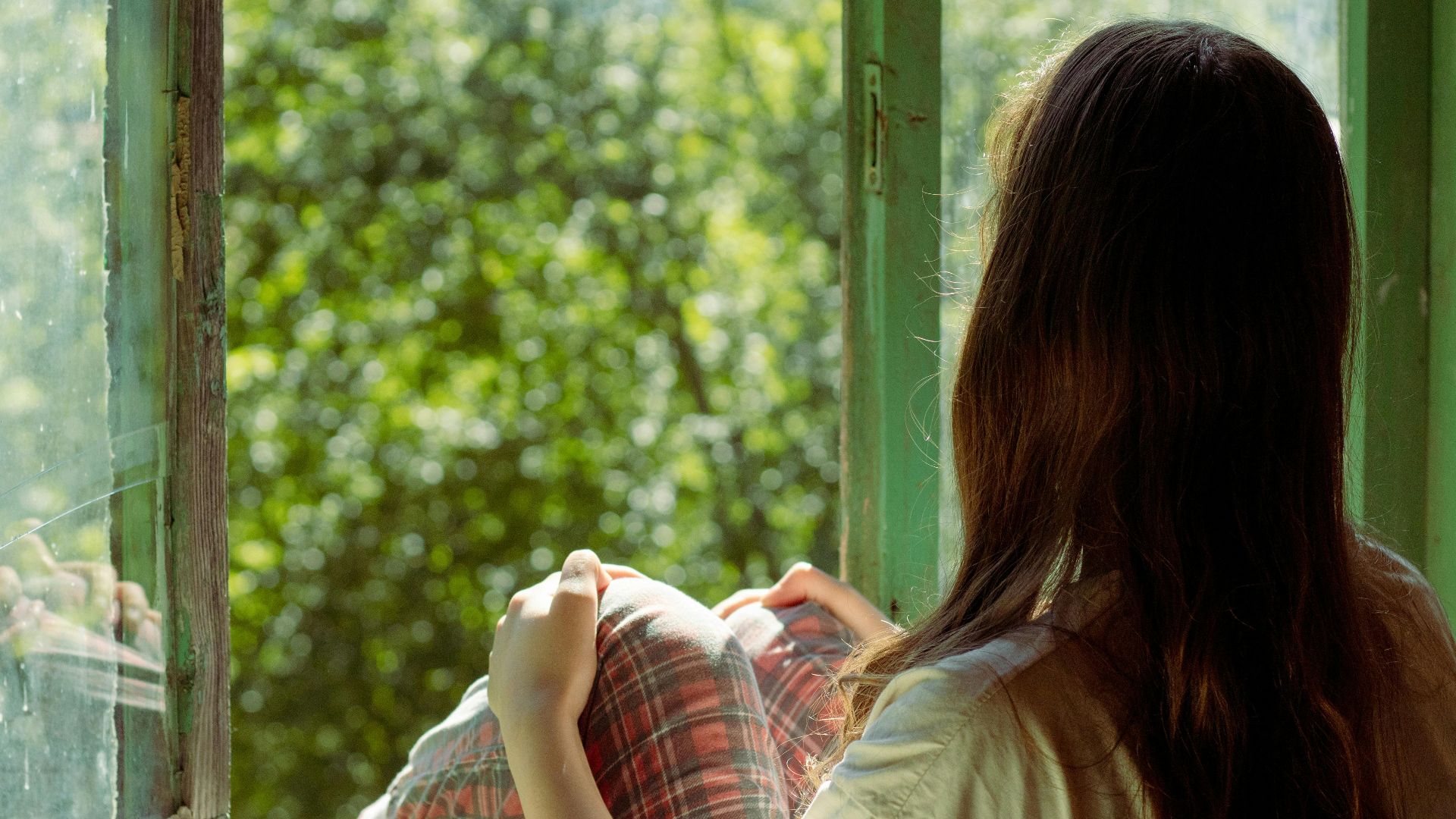 Woman sitting by an open window enjoying the view of lush greenery outside. Perfect for themes of relaxation and solitude.