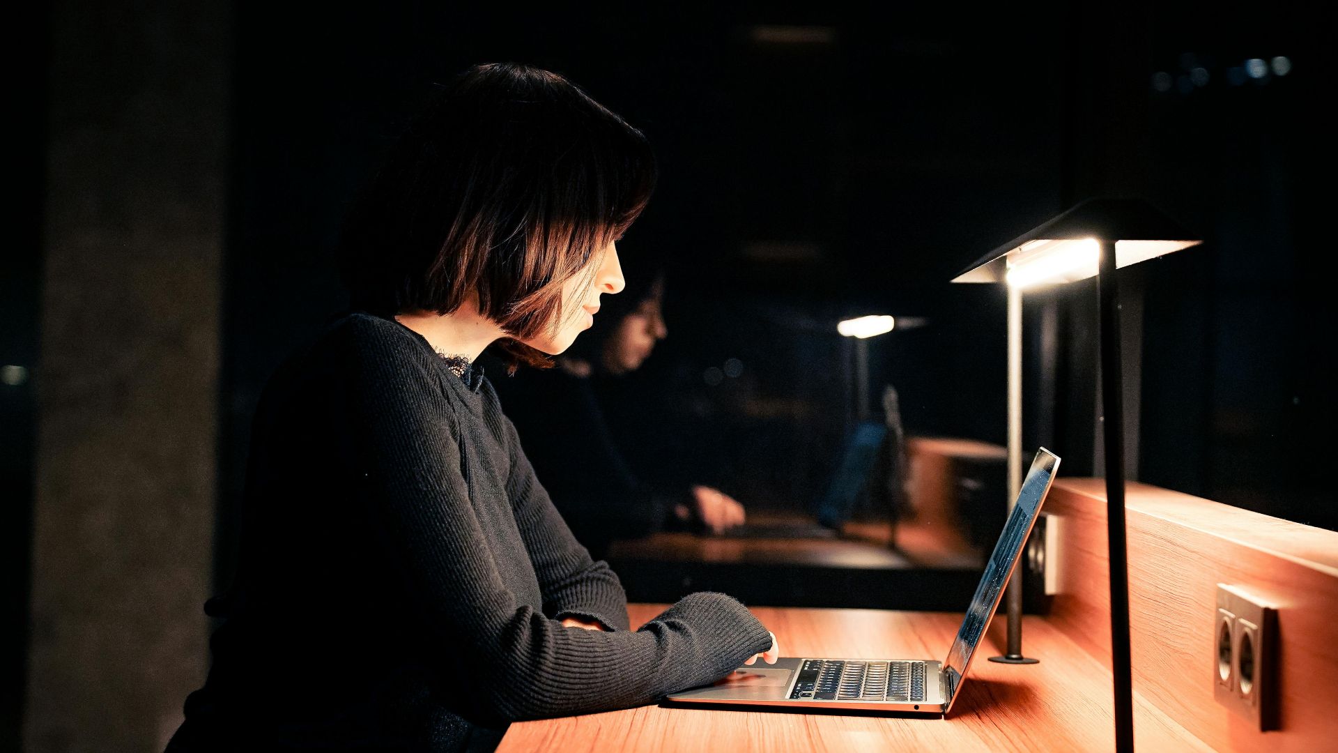 A woman focused on her laptop at a desk with dim lighting, working late at night.